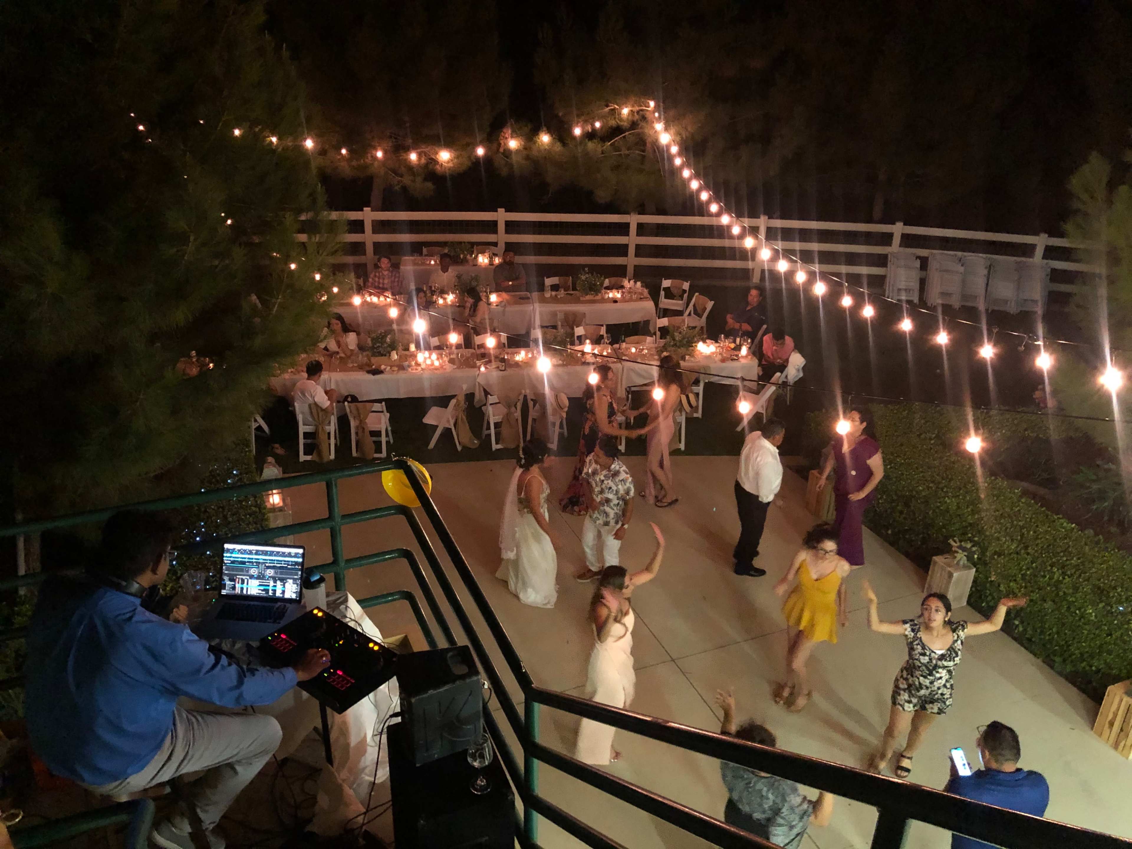 A DJ mixes music while a group of people dance under string lights at an outdoor wedding reception.