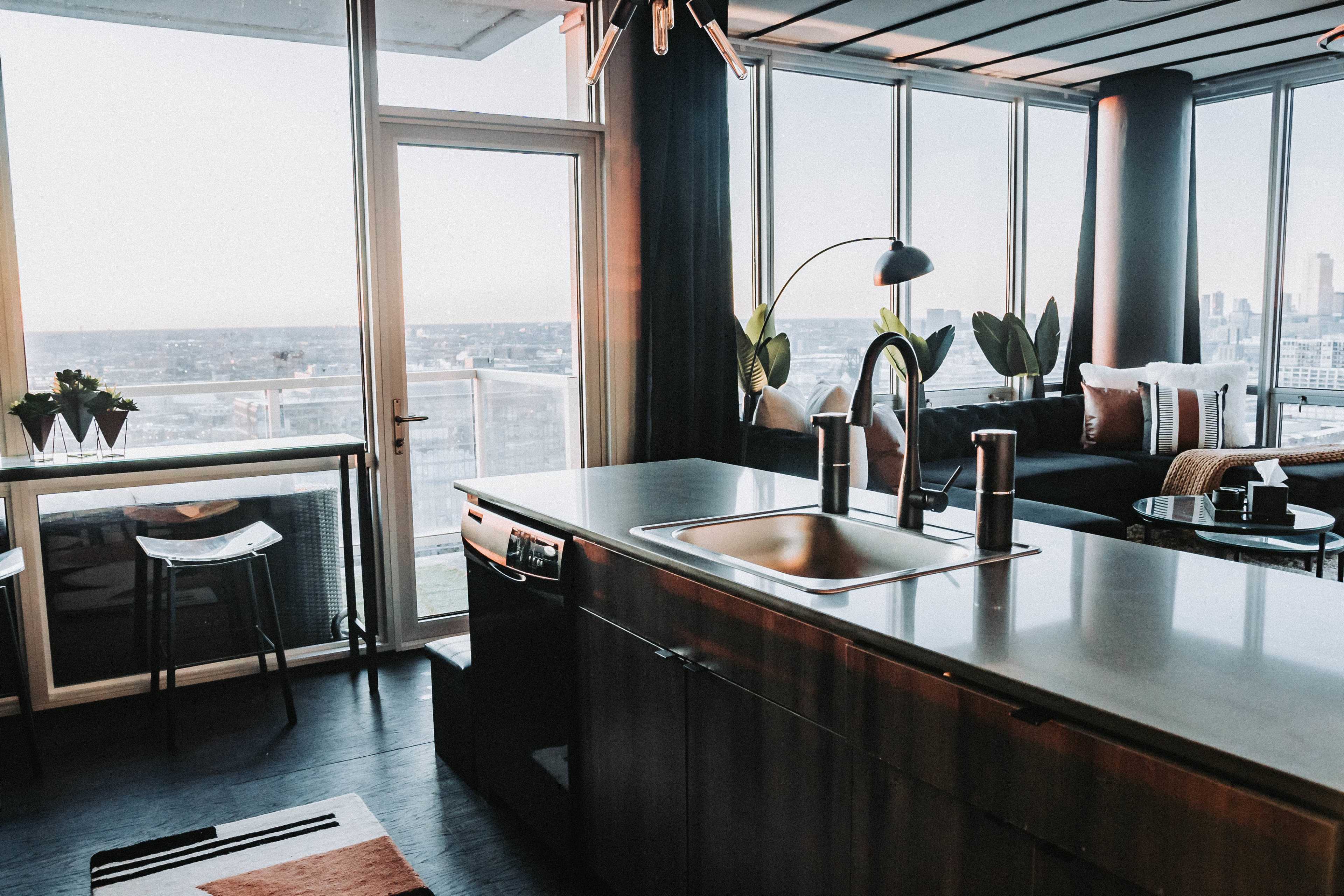 A modern kitchen with a sink and countertop overlooks a bright living area featuring large windows with a city view.