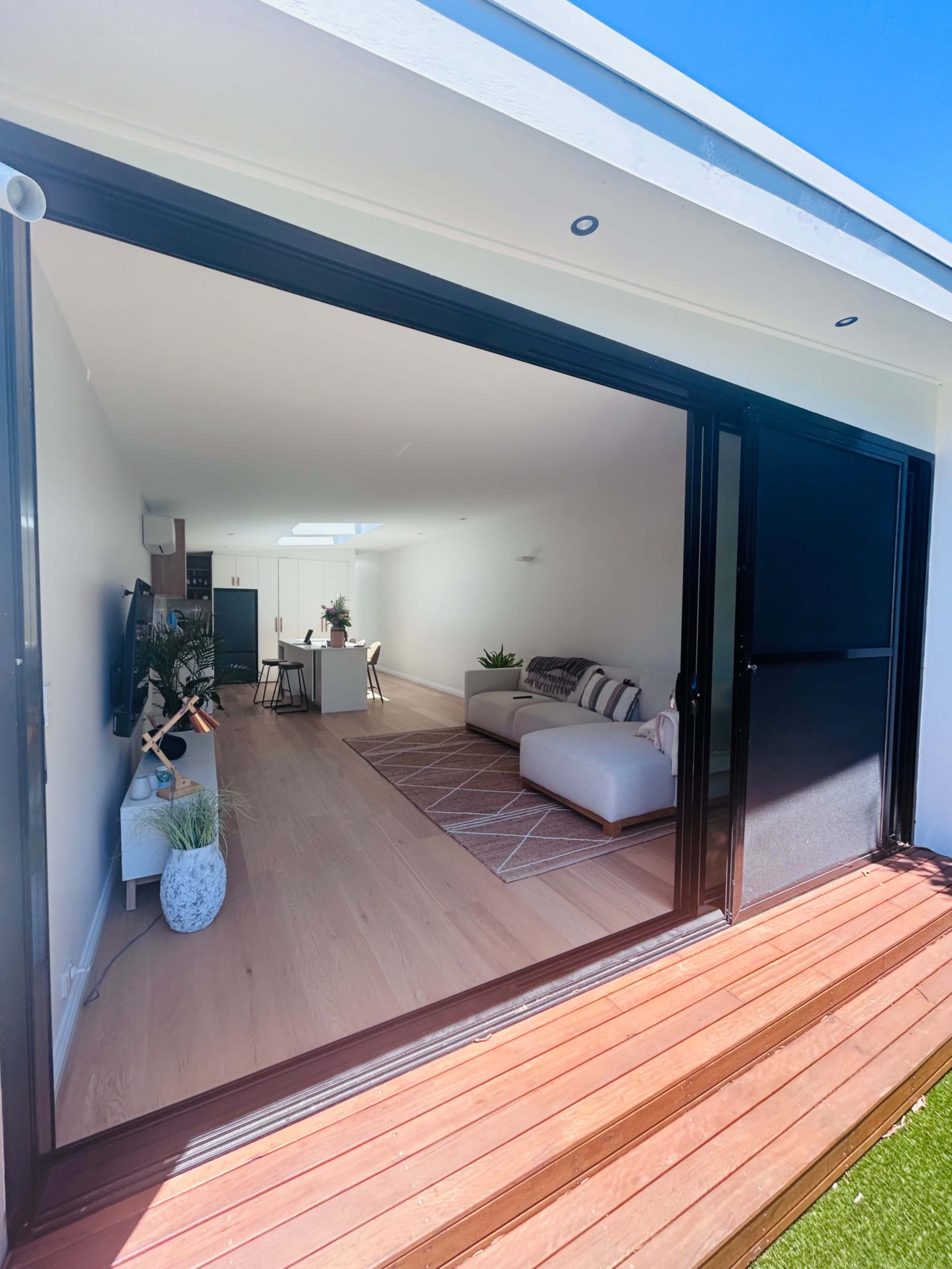 The image shows a modern living space with an open layout, featuring large sliding glass doors leading to a wooden deck.