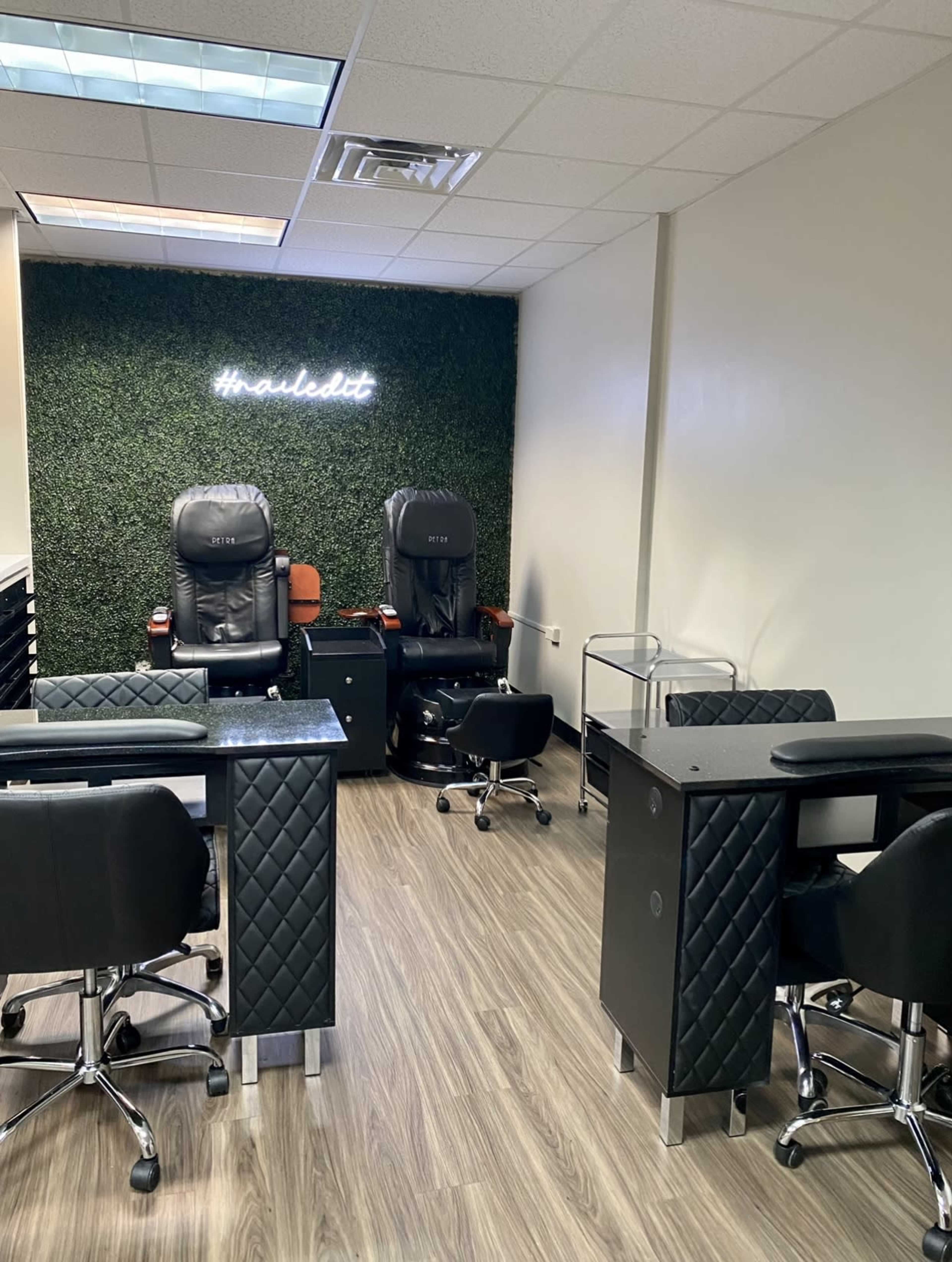 A nail salon interior with two pedicure chairs, nail stations, and a greenery wall feature.