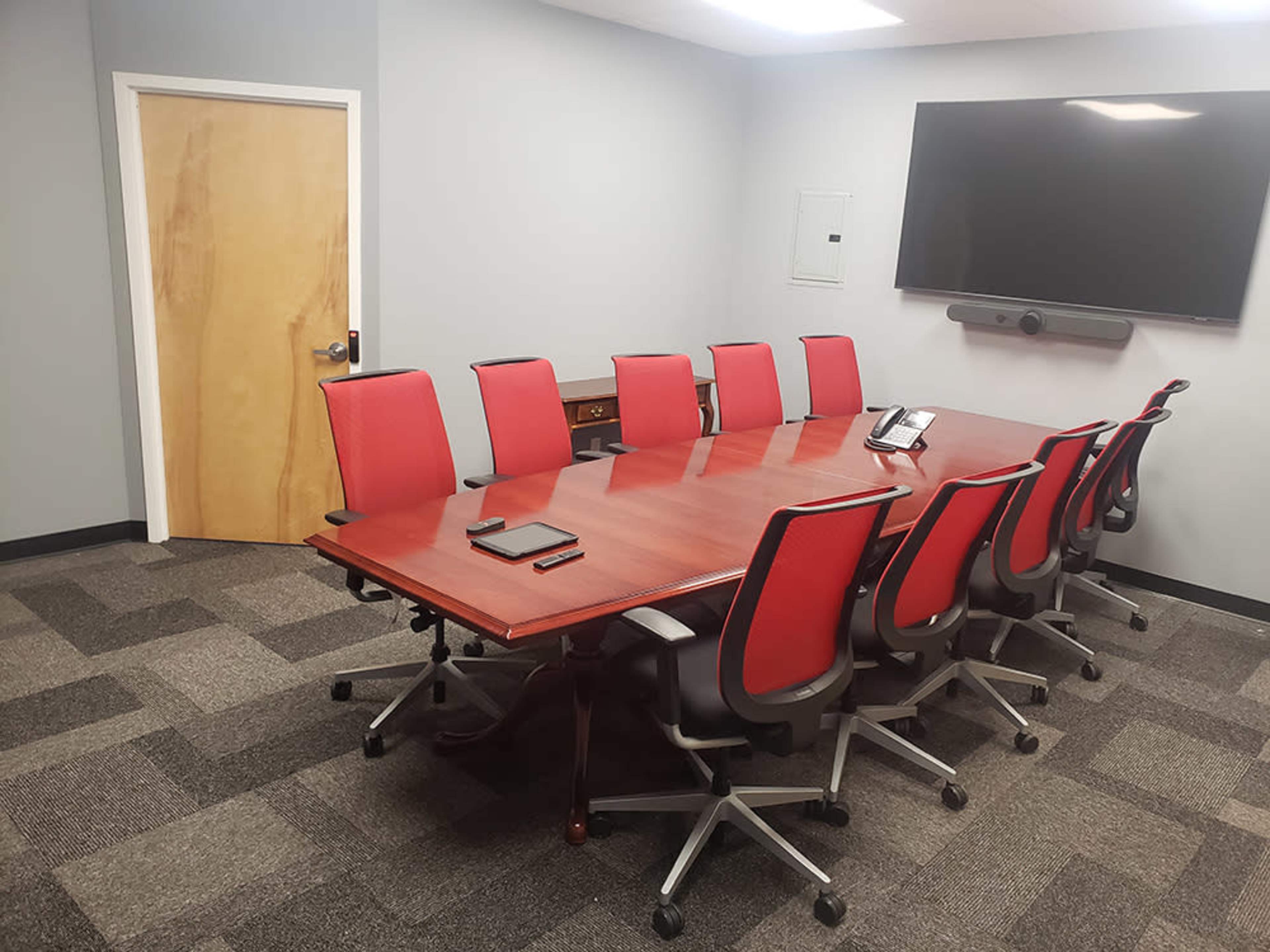A conference room features a long wooden table surrounded by red office chairs, with a large screen mounted on the wall.