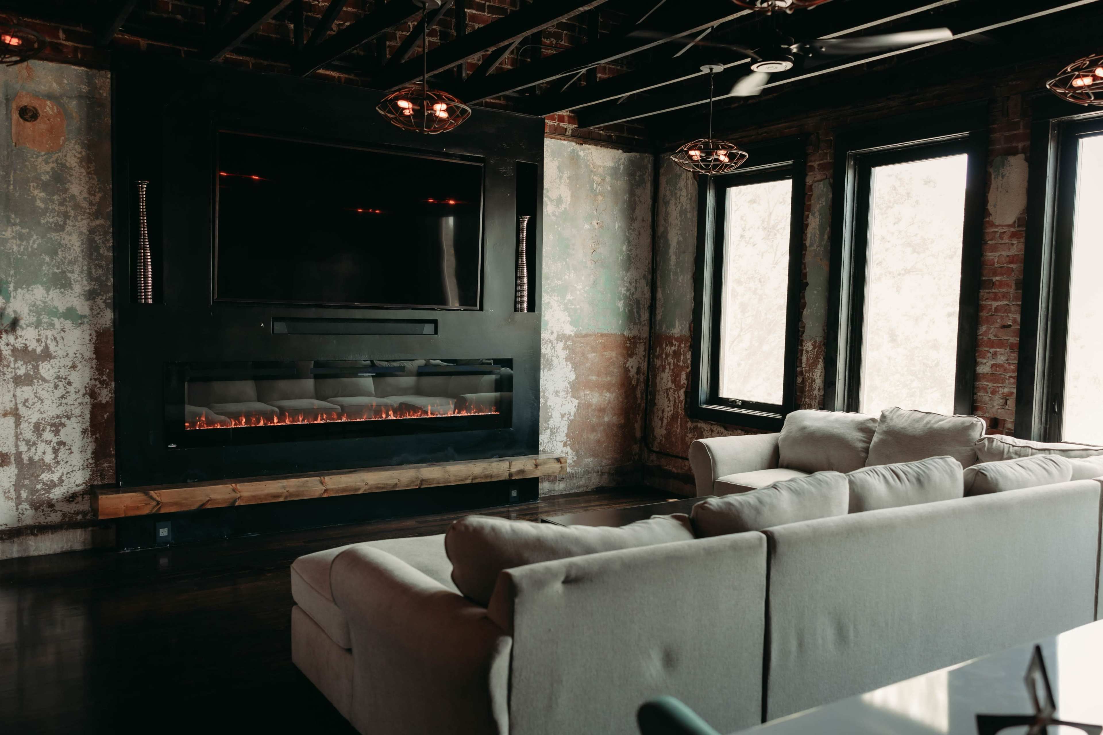 A modern living room features a large sectional sofa facing an electric fireplace mounted beneath a flat-screen TV, with large windows allowing natural light to enter the space.