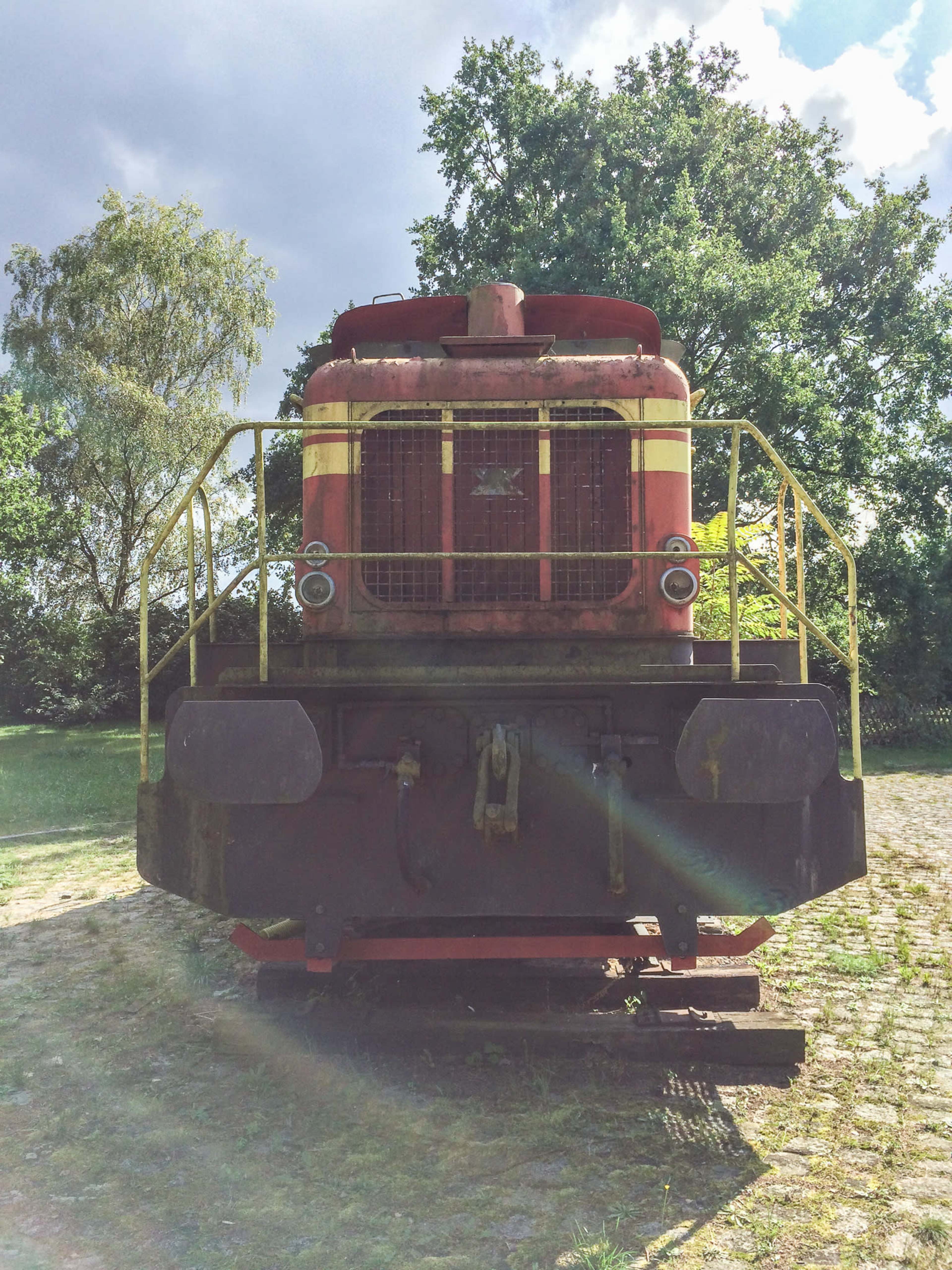 A vintage red locomotive sits on a cobblestone path surrounded by greenery.