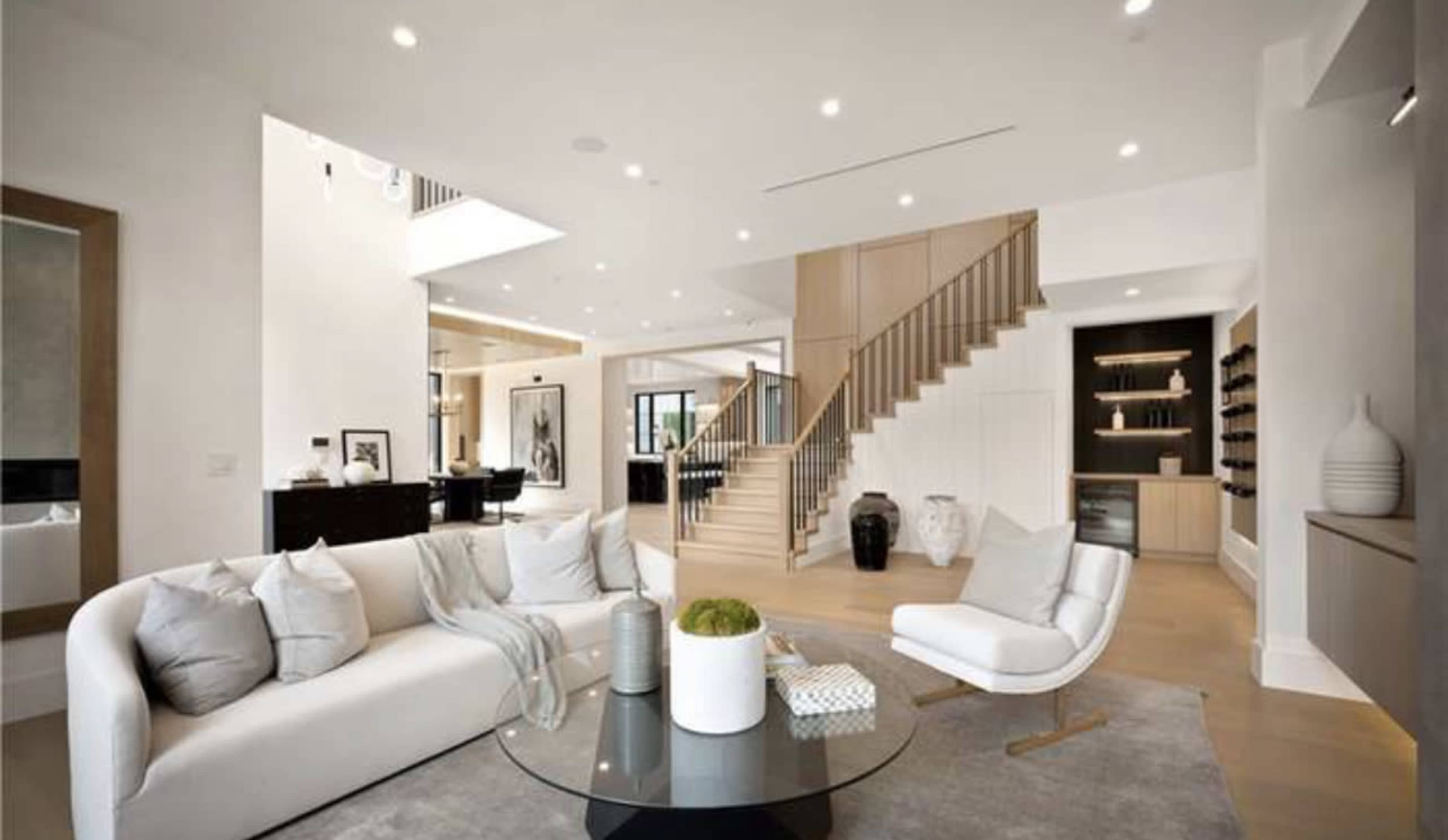 The room features a modern living space with a white sectional sofa, a circular glass table, and a wooden staircase leading to an upper level.