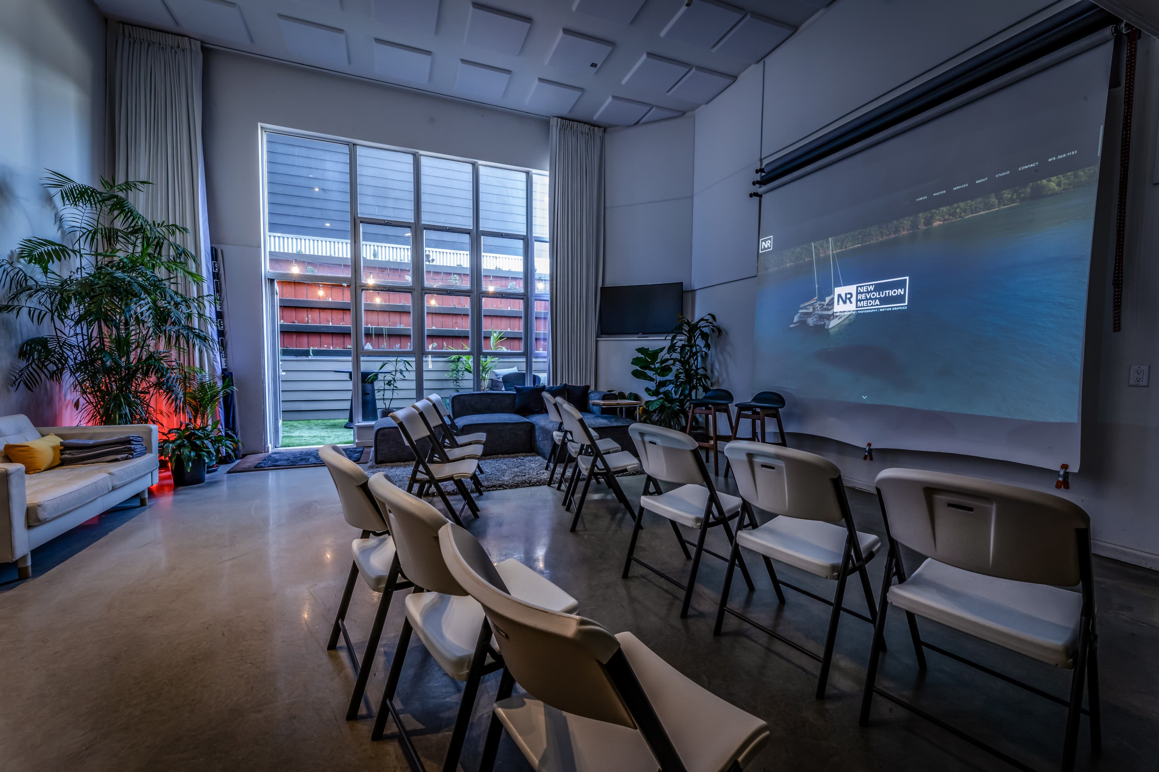 A modern room contains rows of white chairs facing a projection screen, with large windows revealing an outdoor space.