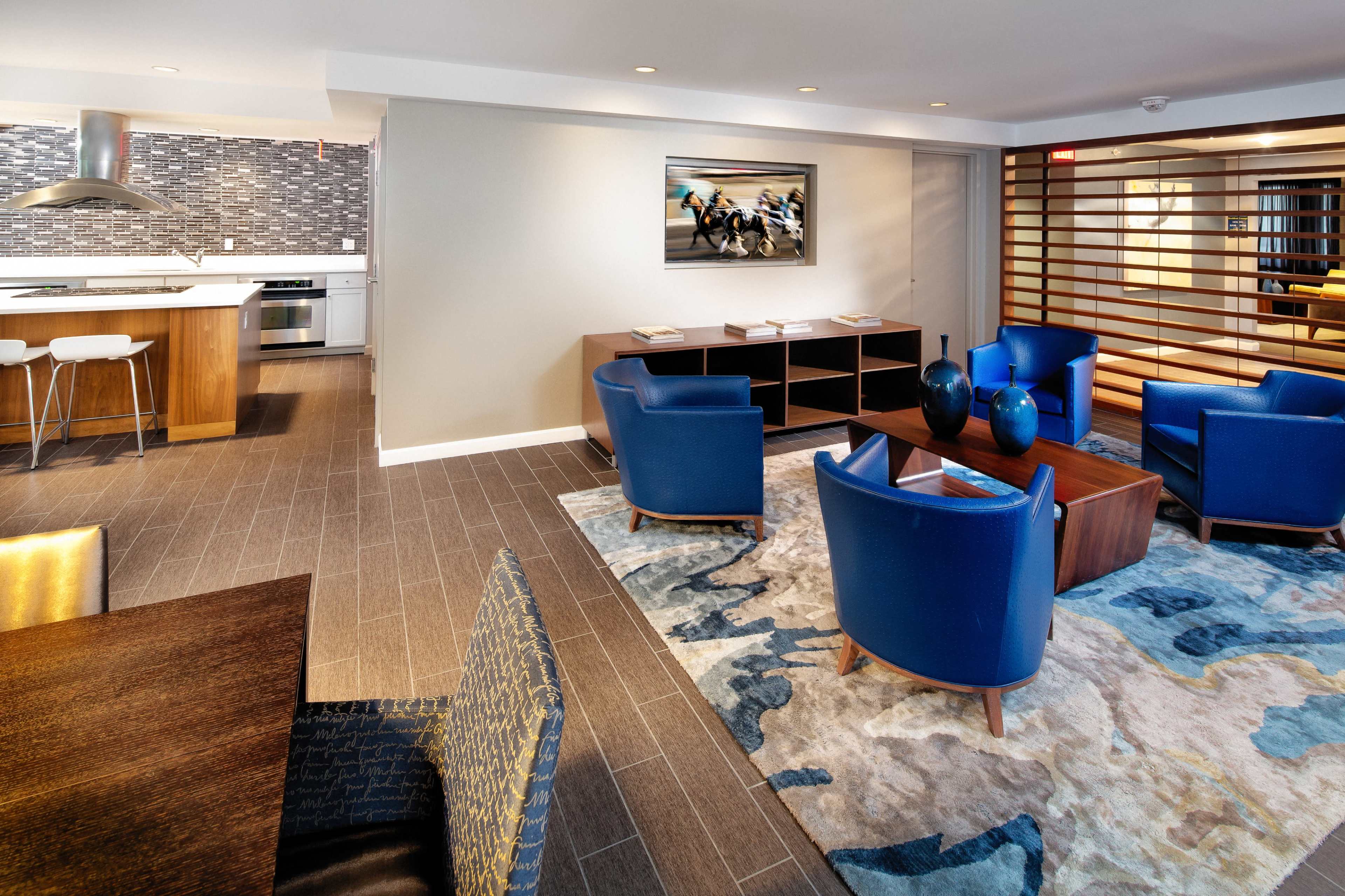 A modern living area with blue chairs, a wooden coffee table, a patterned rug, and a kitchen area in the background featuring a sleek design.