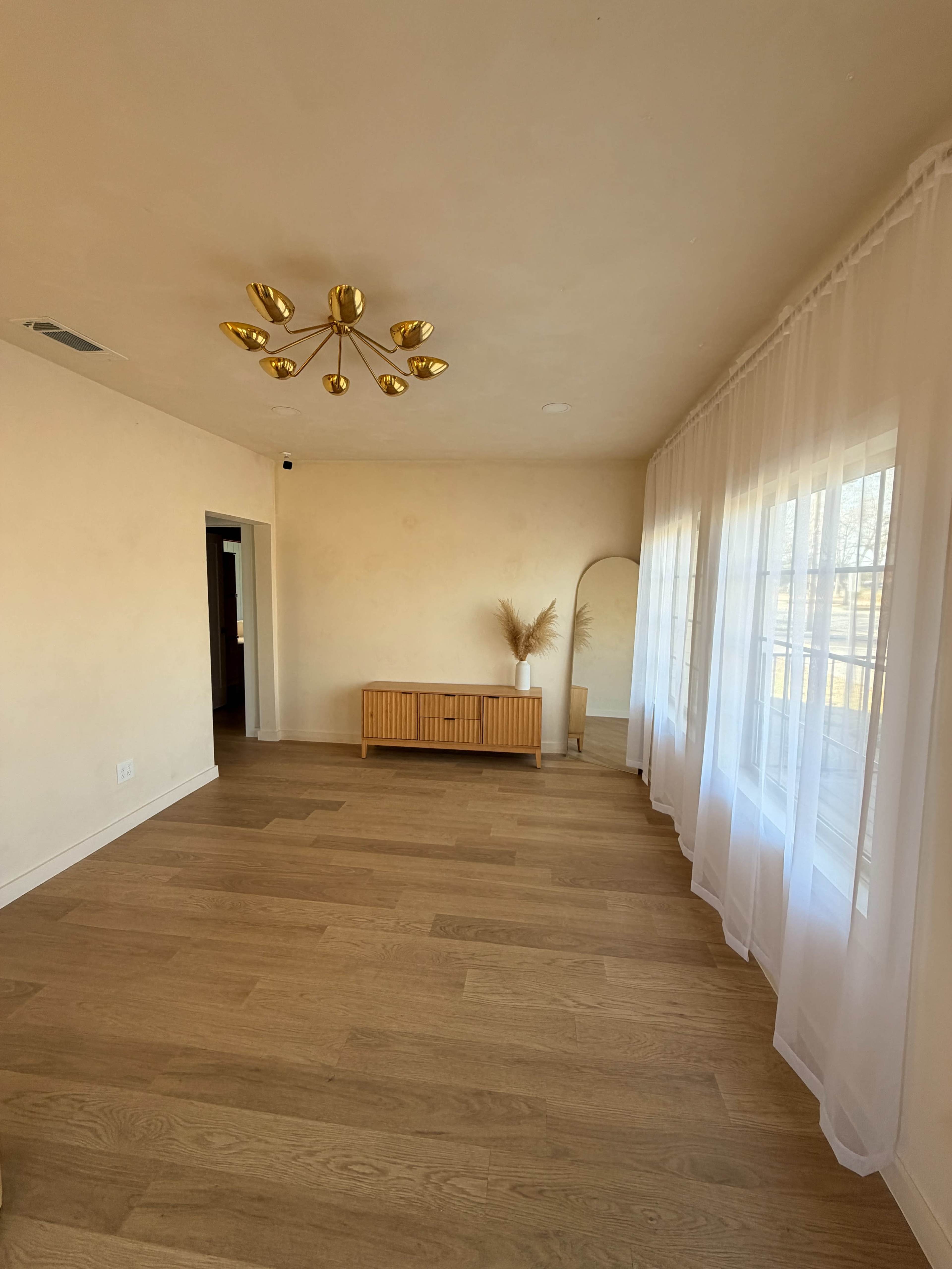 The image shows a minimalist living room with light-colored walls, wooden flooring, a wooden sideboard, a floor mirror, and large sheer curtains covering a window.