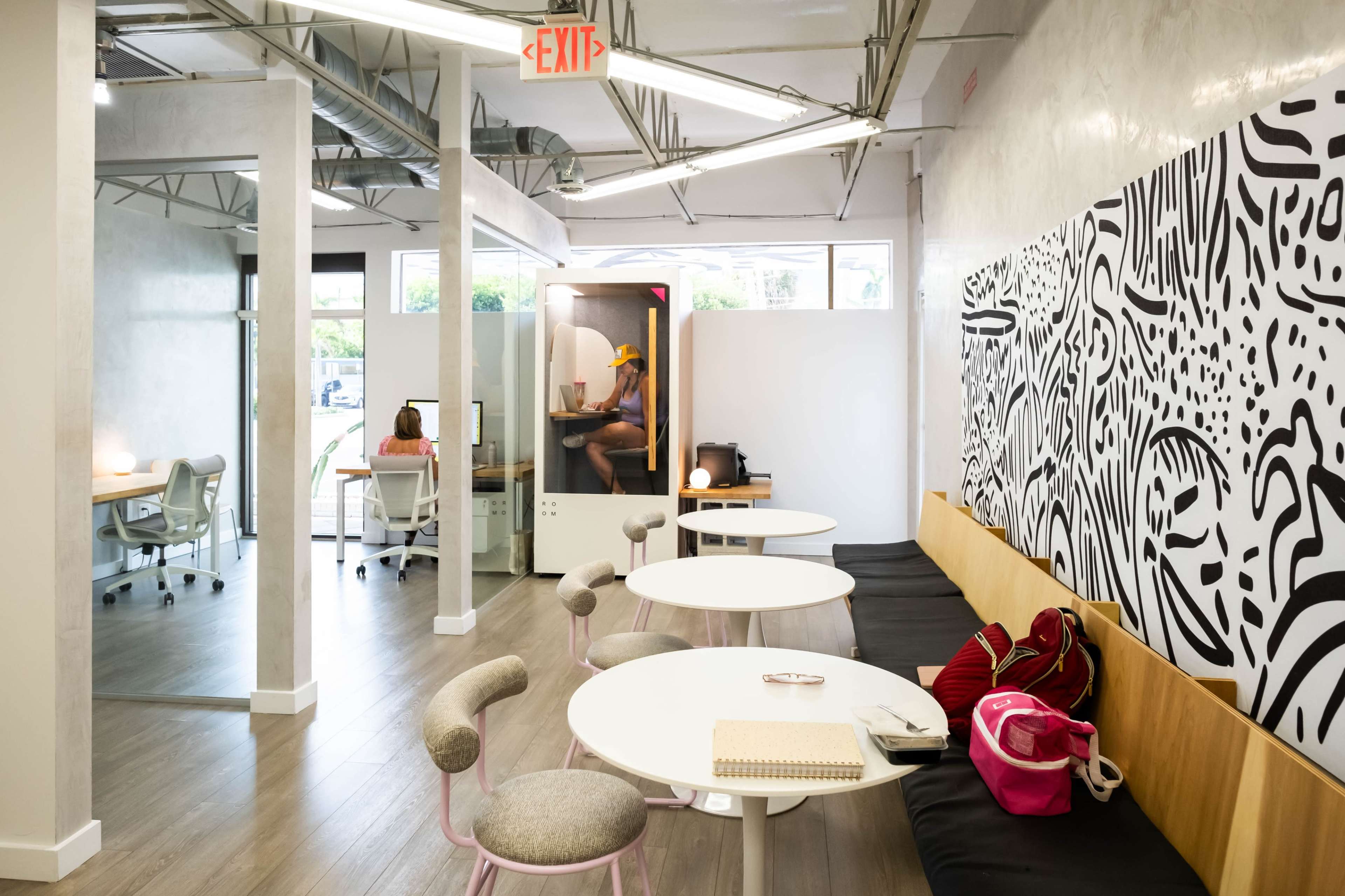 The image shows a modern office space with several workstations, a seating area, and a soundproof pod in the corner.