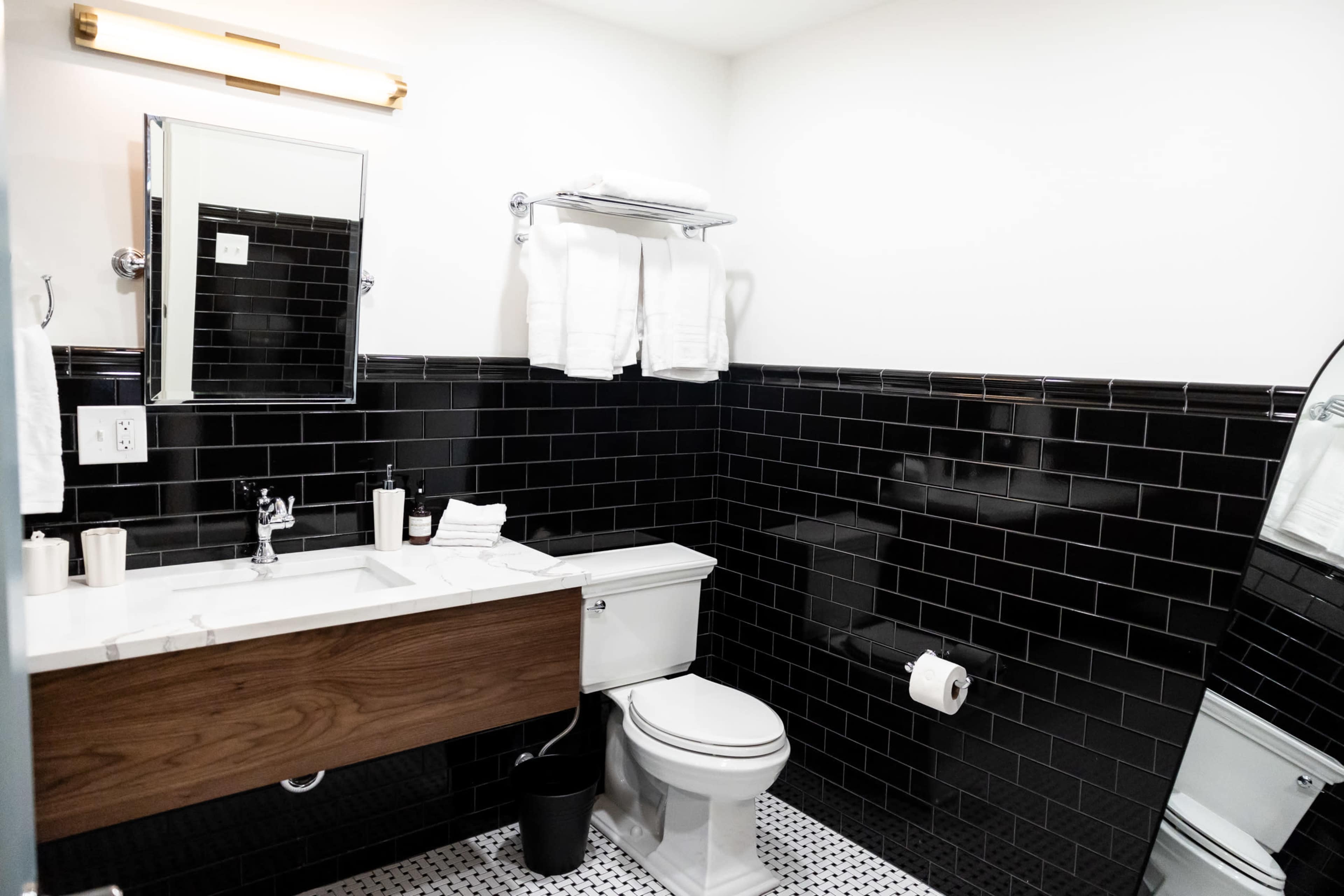 The image shows a modern bathroom featuring black tile walls, a wooden vanity with a sink, a toilet, and neatly arranged towels.