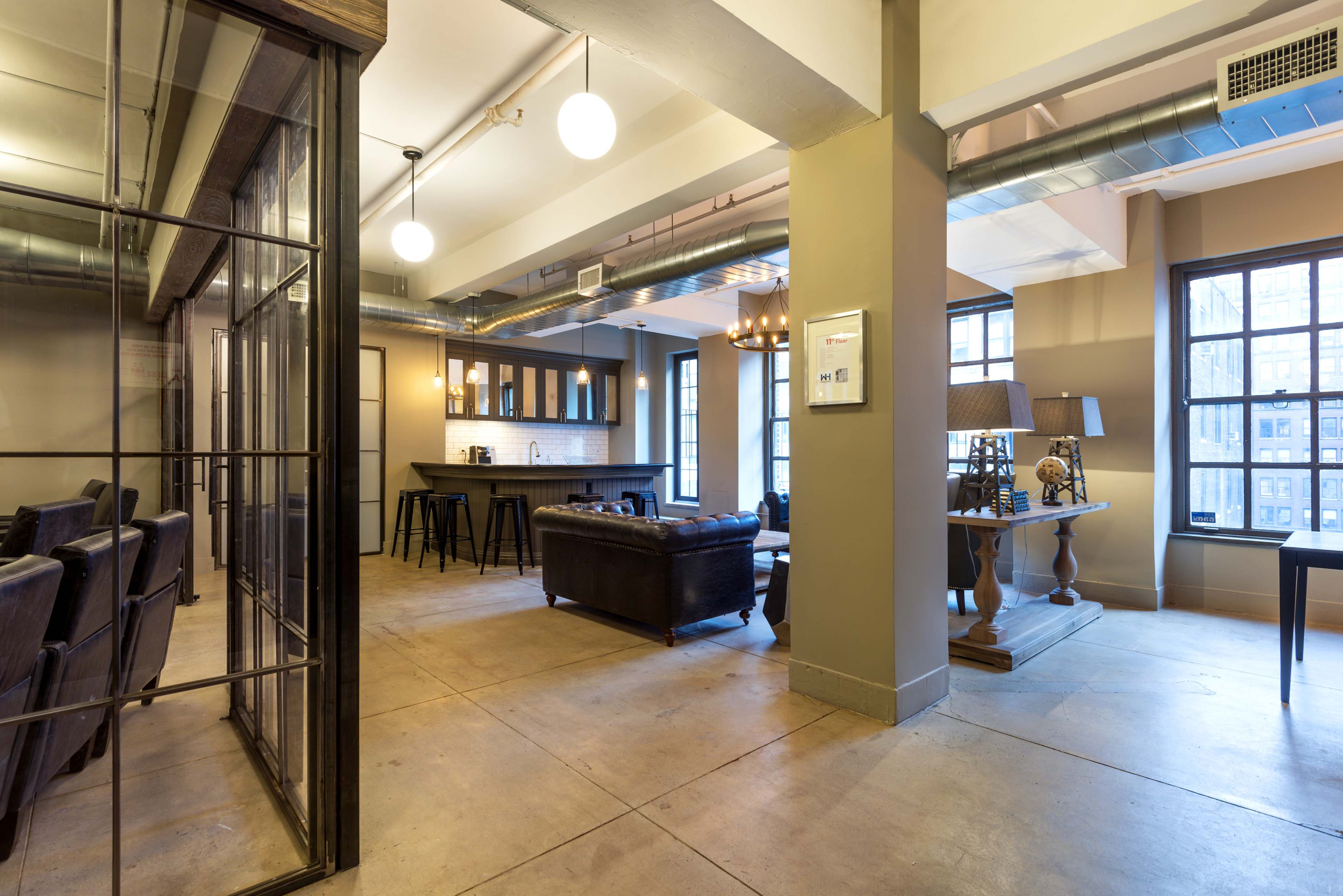 A spacious, modern interior featuring a living area with a leather sofa, a kitchen area with bar stools, and large windows allowing natural light to enter.