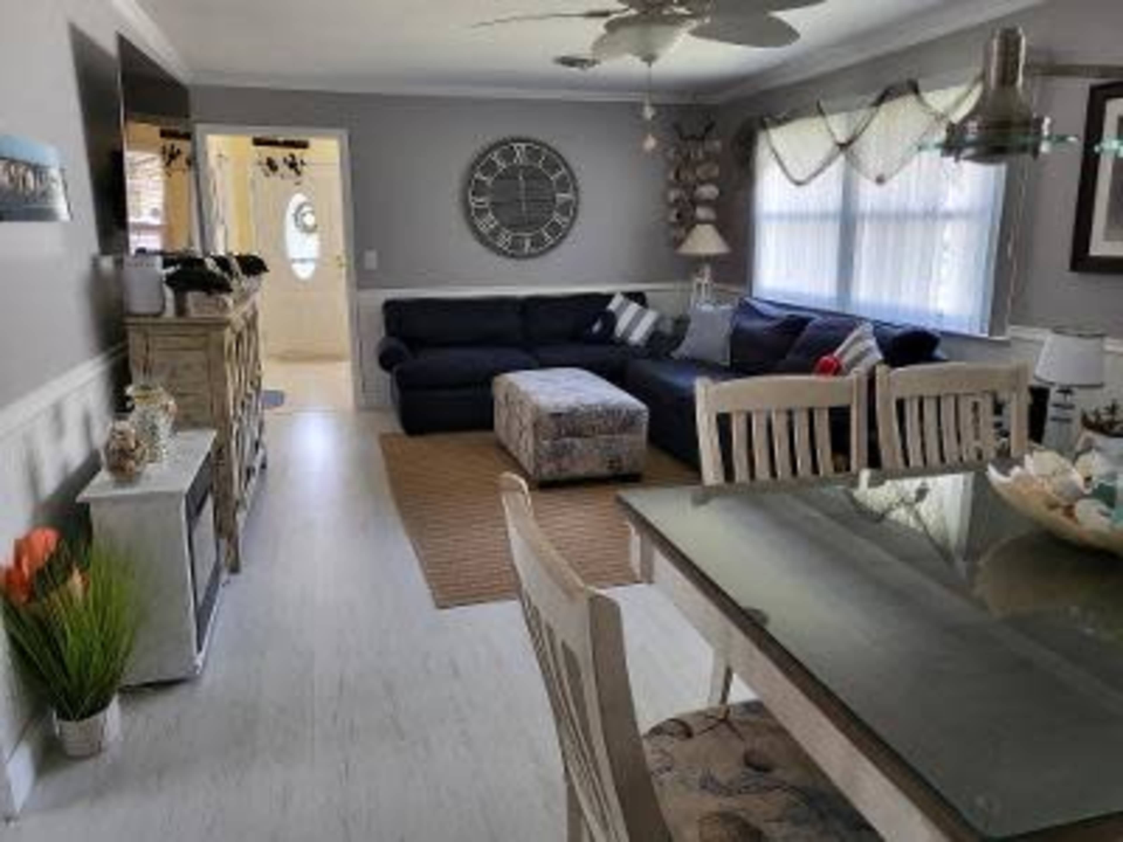 A living room and dining area feature a dark sectional sofa, a square ottoman, a dining table with chairs, and a wall clock.