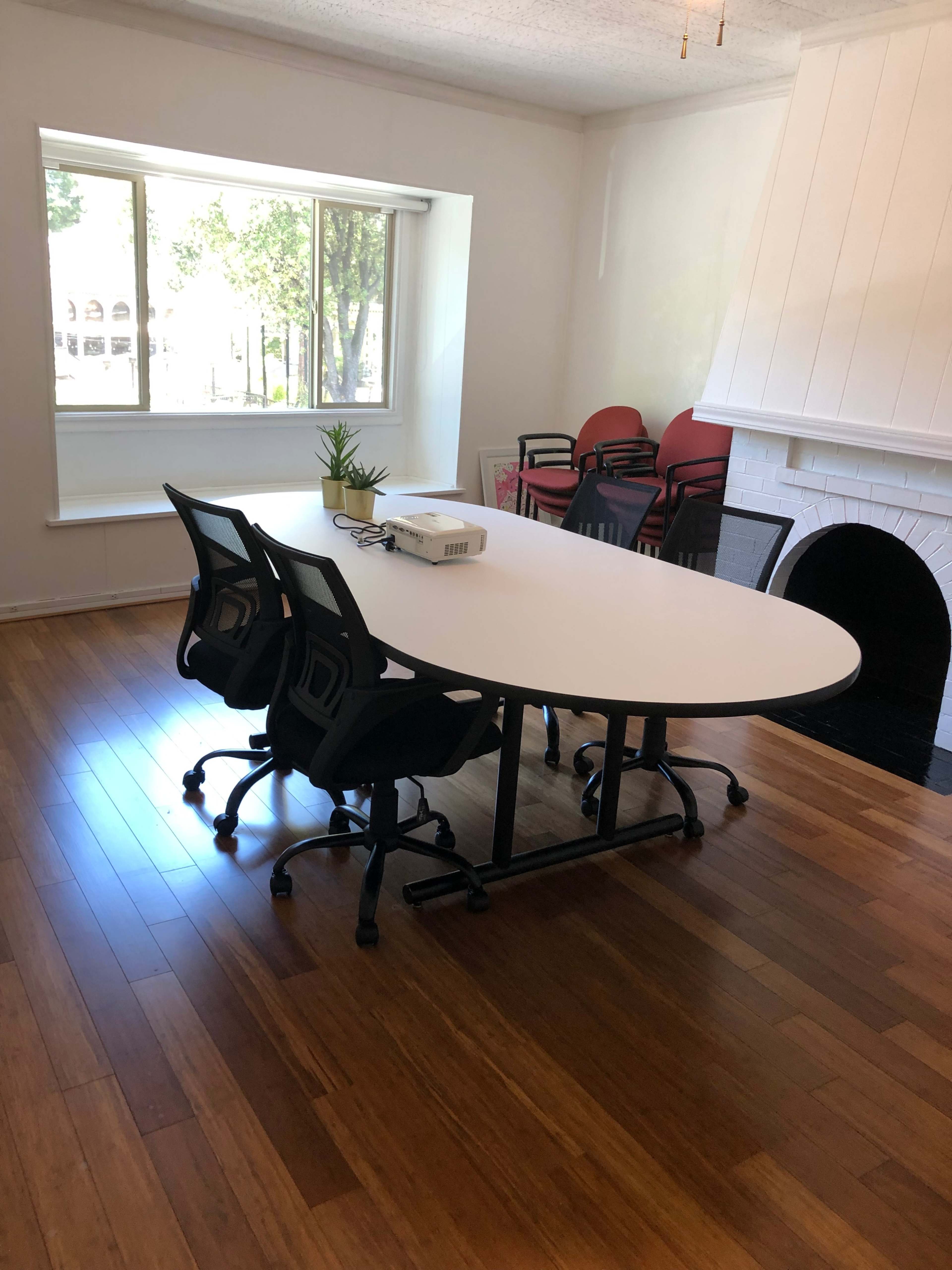 A modern conference room features an oval table with four rolling chairs, a projector on the table, and a large window allowing natural light to enter.