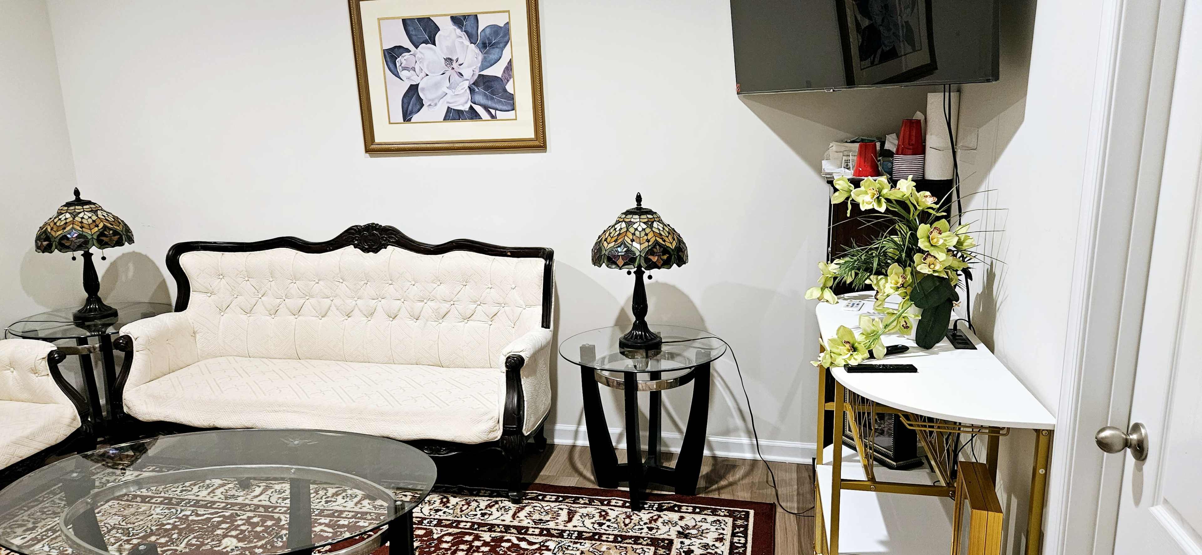 The image shows a living room with a white tufted sofa, two decorative lamps on glass side tables, a large floral wall art piece, and a corner table displaying a plant arrangement and a television.