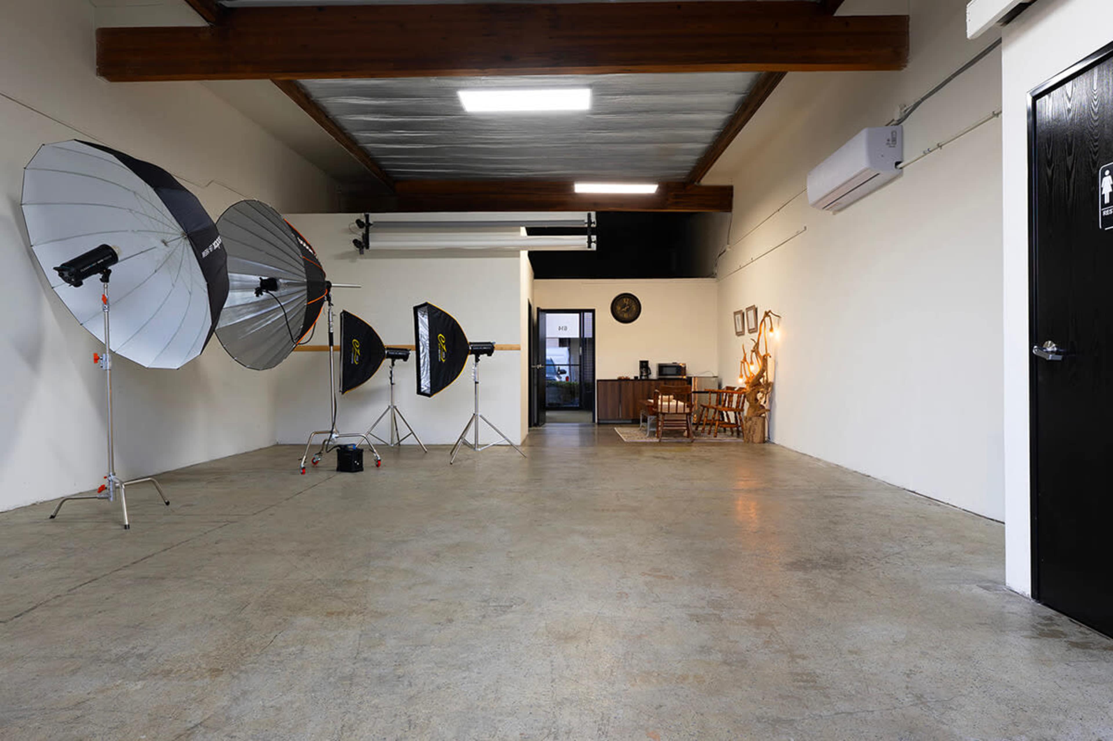 The image shows a spacious, empty studio with large photography reflectors on the left and a doorway leading to a kitchen area in the background.