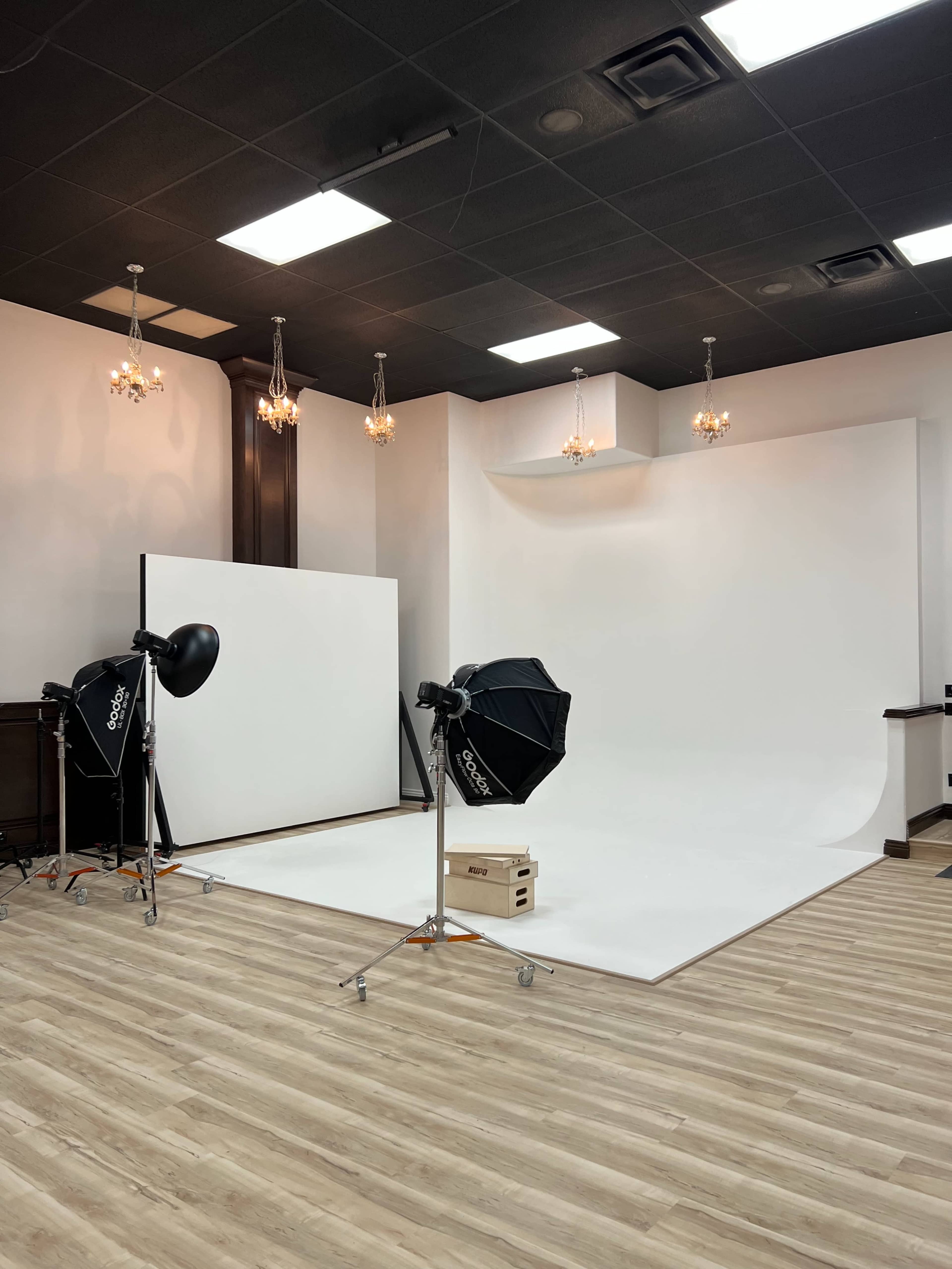 The image shows a photography studio with a white backdrop, lighting equipment, and wooden flooring.