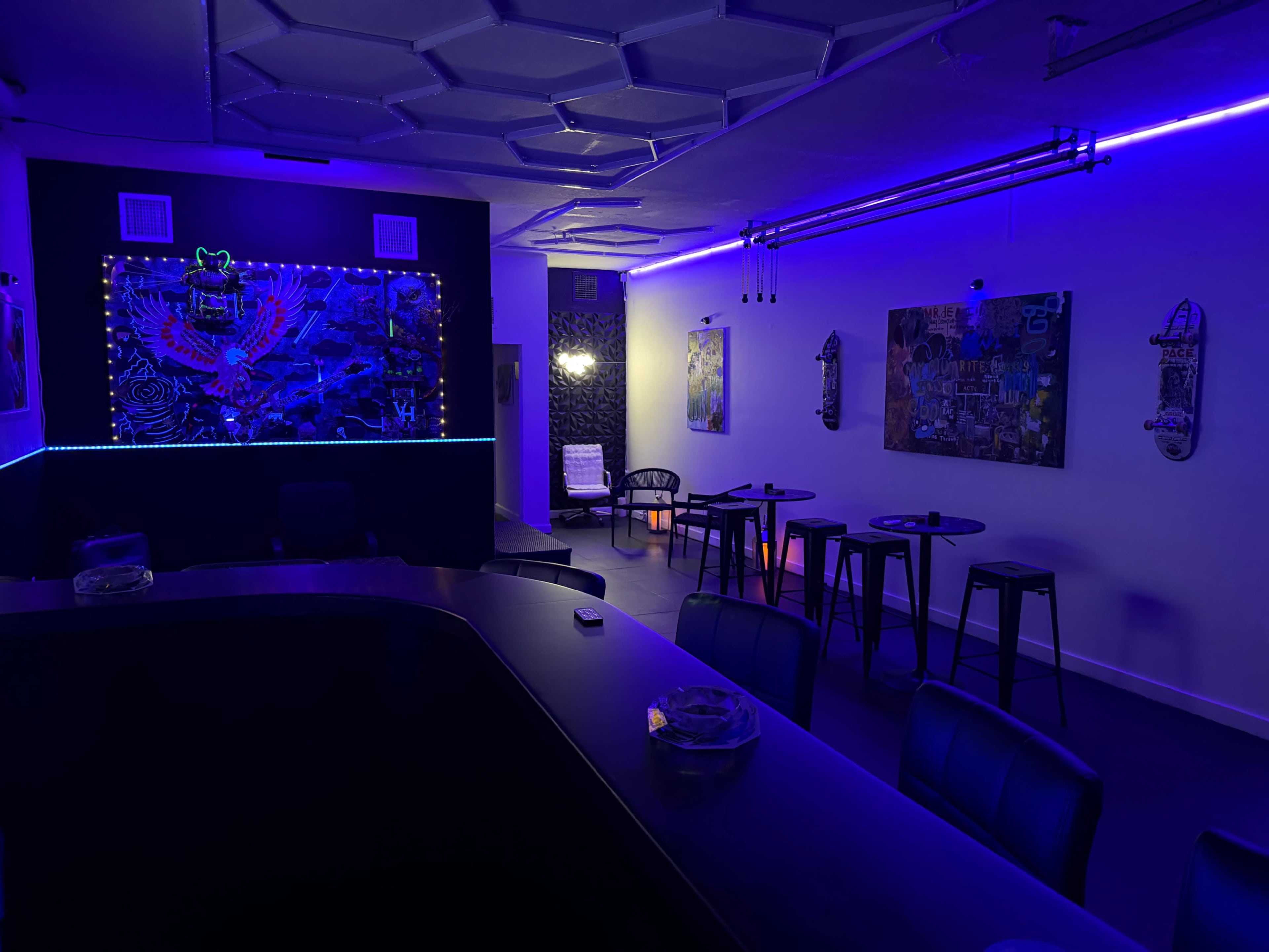The image shows a dimly lit lounge area featuring a bar counter, high tables with stools, and neon blue lighting accentuating the wall art.