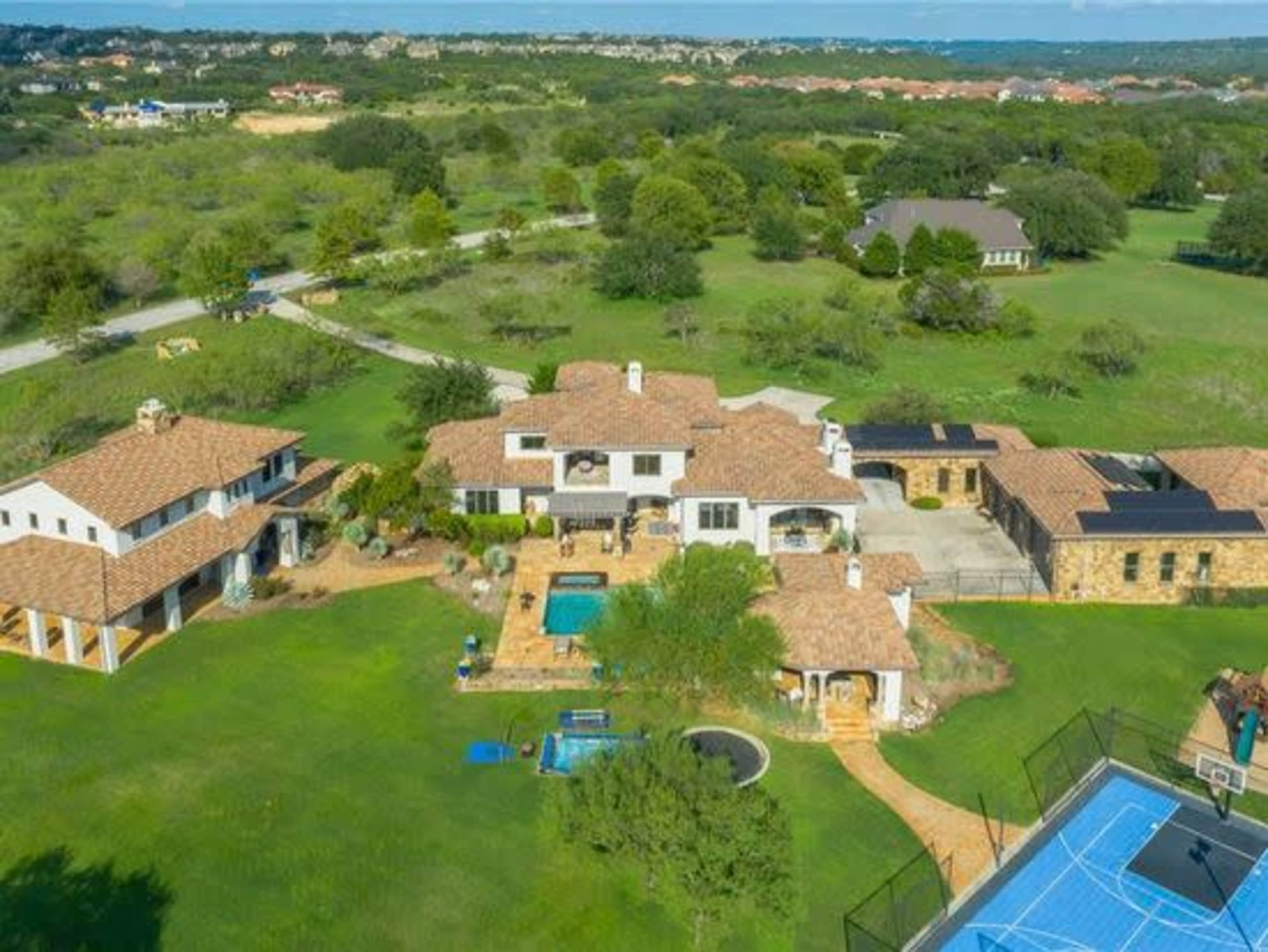 The aerial view shows a sprawling estate with multiple buildings, a swimming pool, a tennis court, and landscaped green lawns.