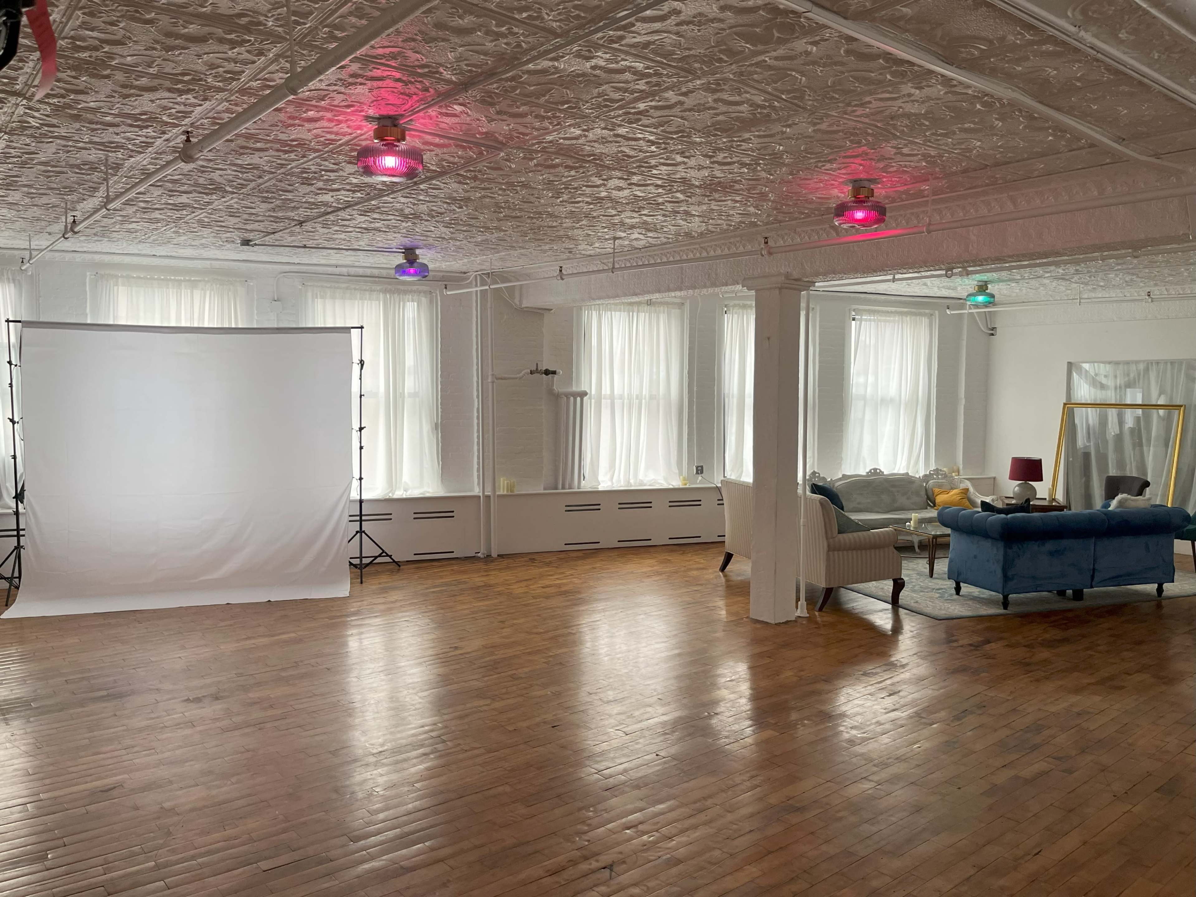 The image shows a spacious, light-filled studio with a white backdrop on one side and a furnished seating area featuring a blue couch and a large mirror on the other.