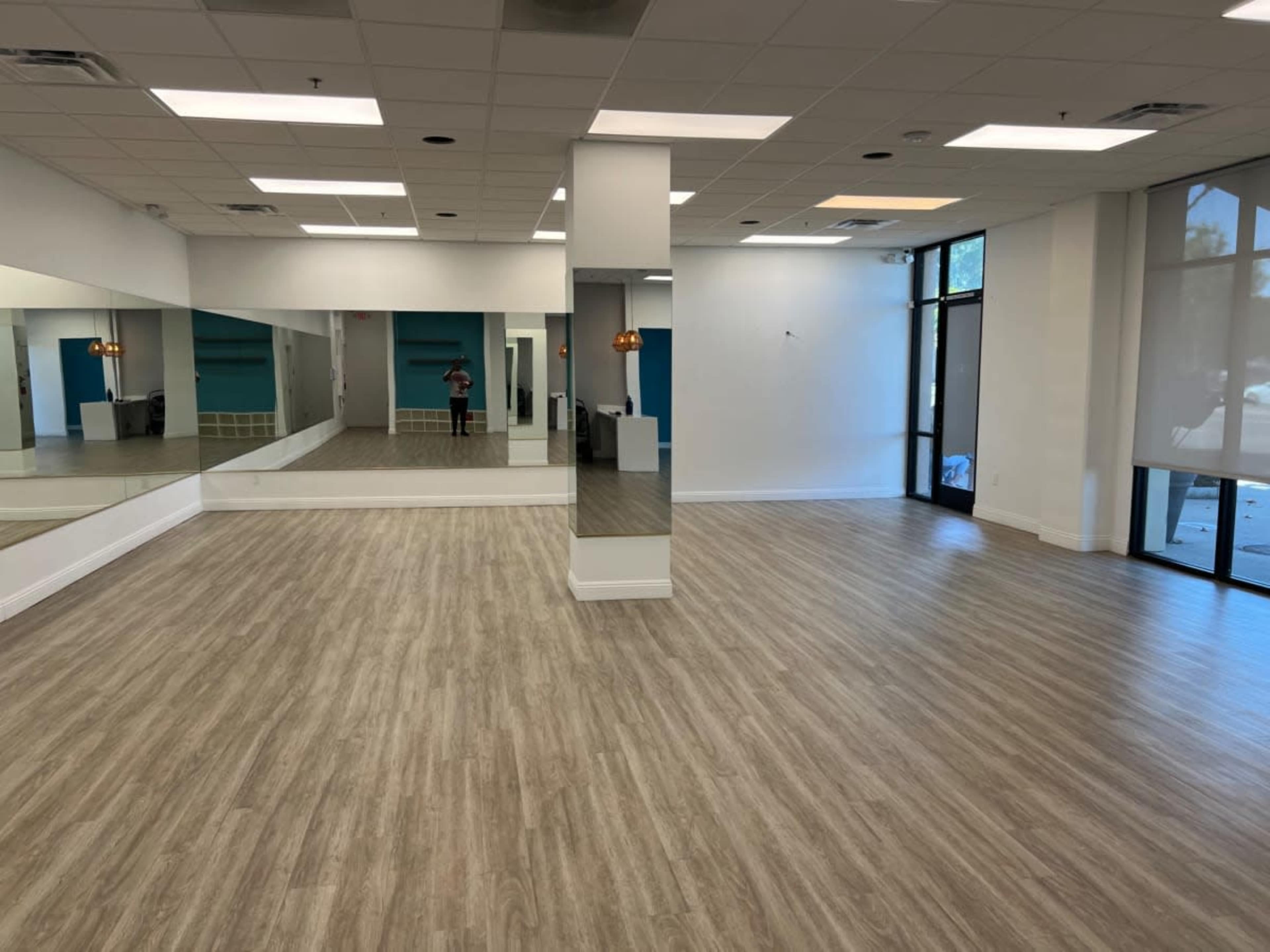 A spacious, brightly lit room with wooden flooring, large mirrors on one wall, and windows allowing natural light.