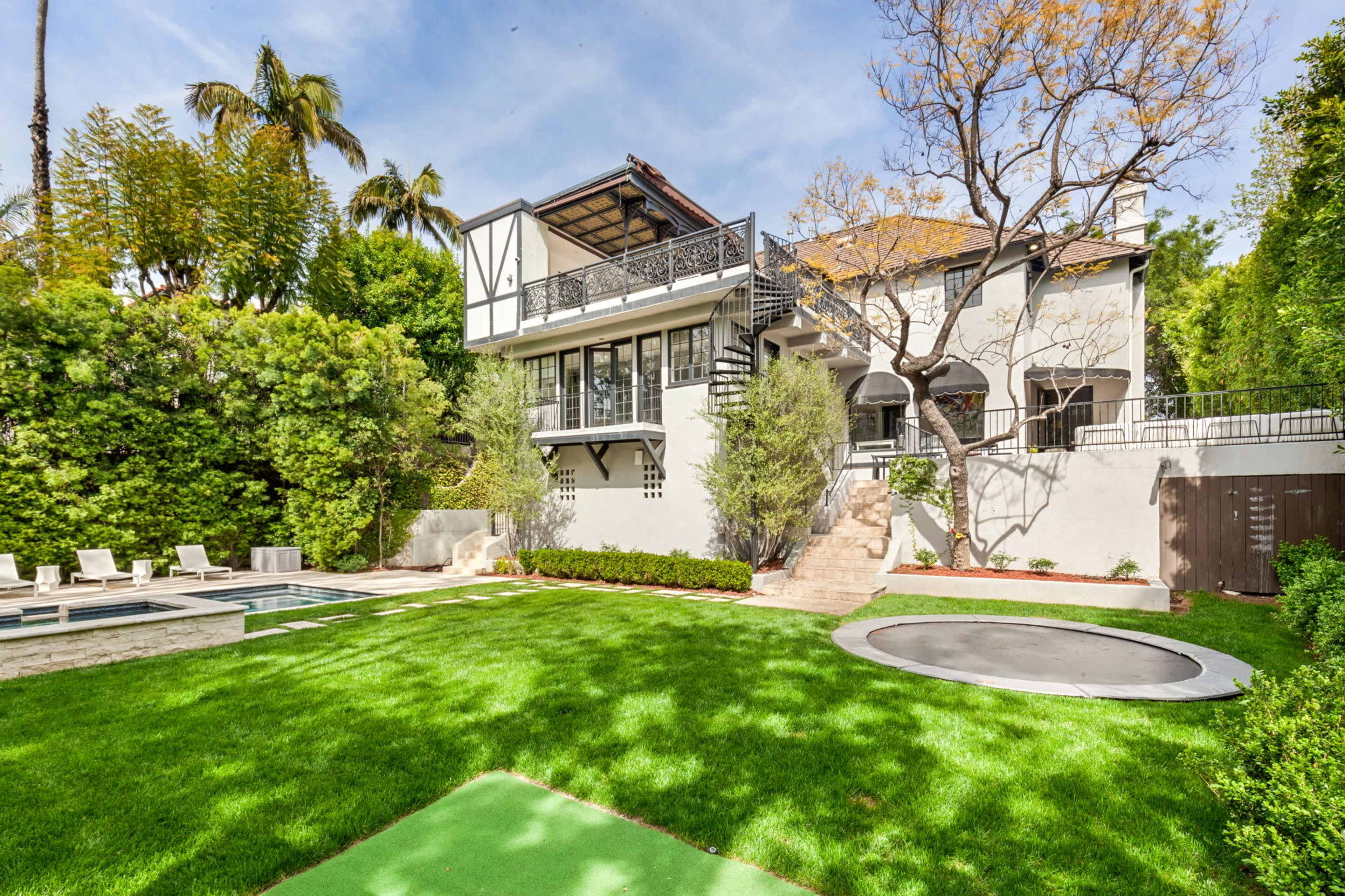 The image shows a spacious house surrounded by greenery, featuring a lawn, a swimming pool, and a trampoline in the backyard.