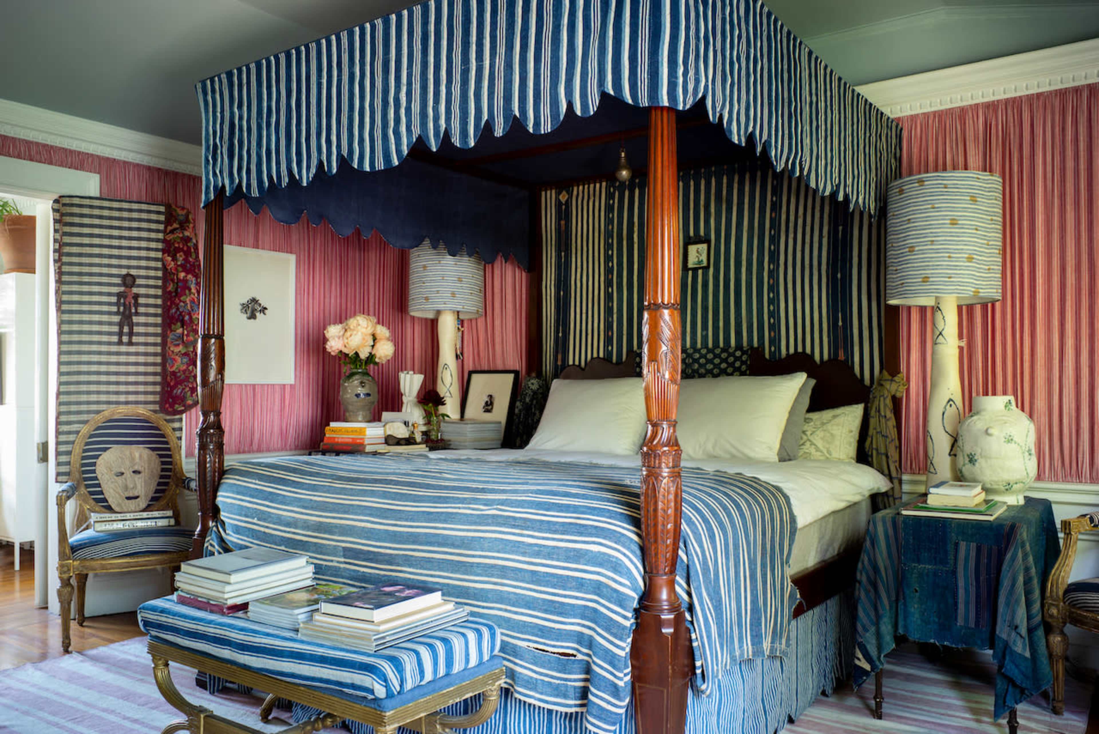 The image shows a bedroom featuring a canopied bed with blue and white striped bedding, complemented by pink walls and various decorative items, including lamps and a chair.