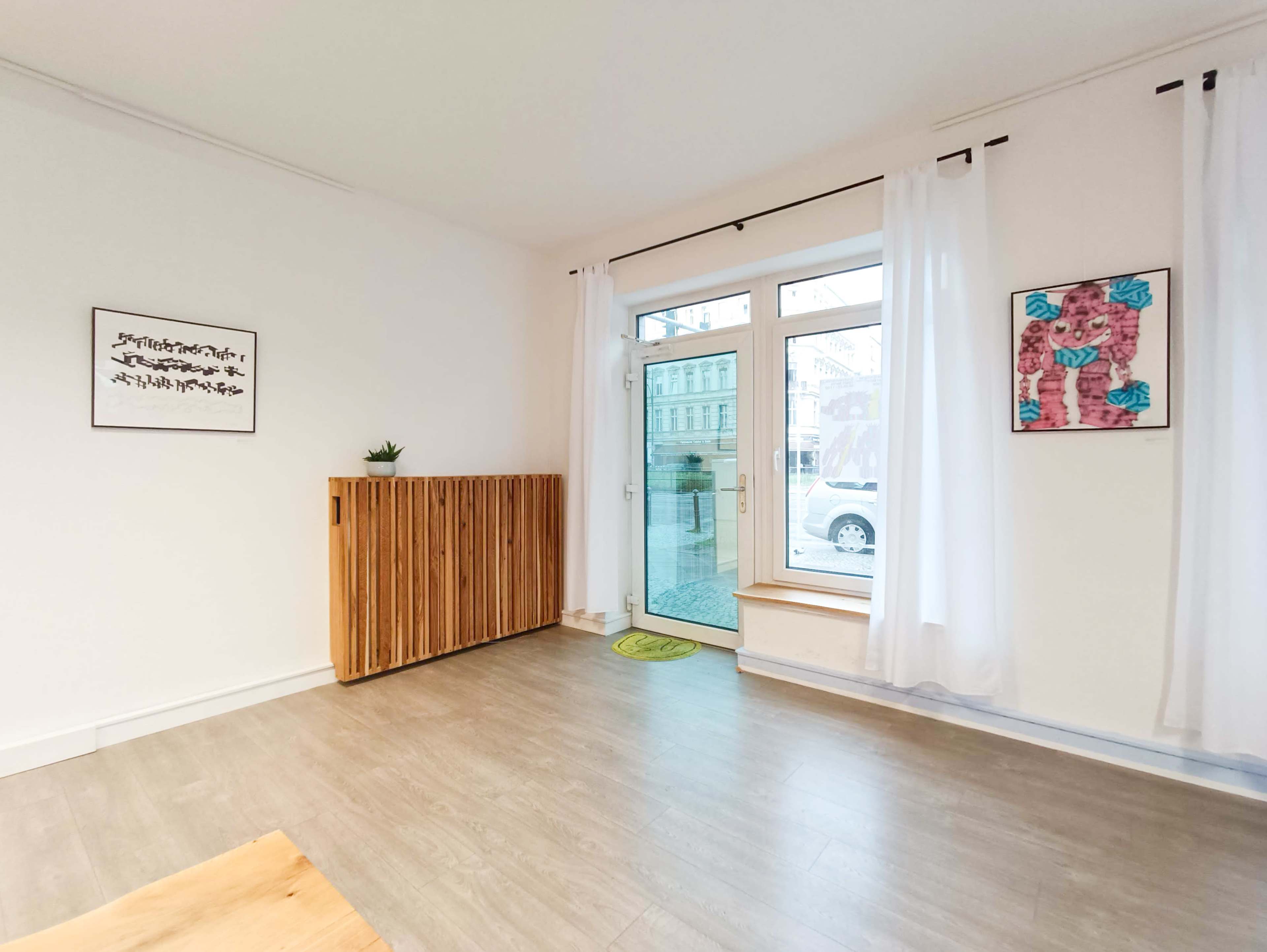 The empty room features large windows with white curtains, a wooden partition, and a small plant on a ledge.