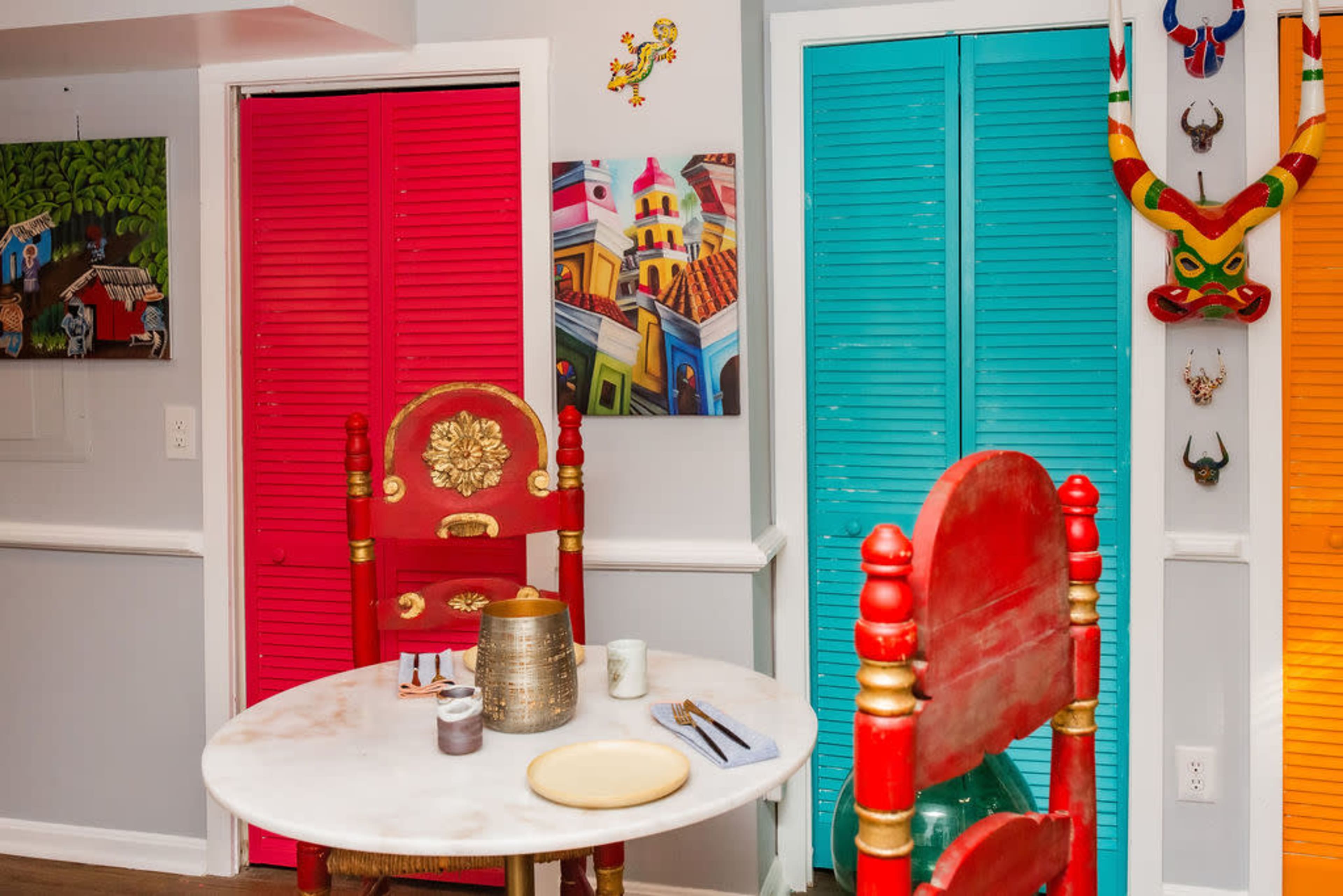 A round table is set in a brightly colored room with red and turquoise shutters and vibrant artwork on the walls.