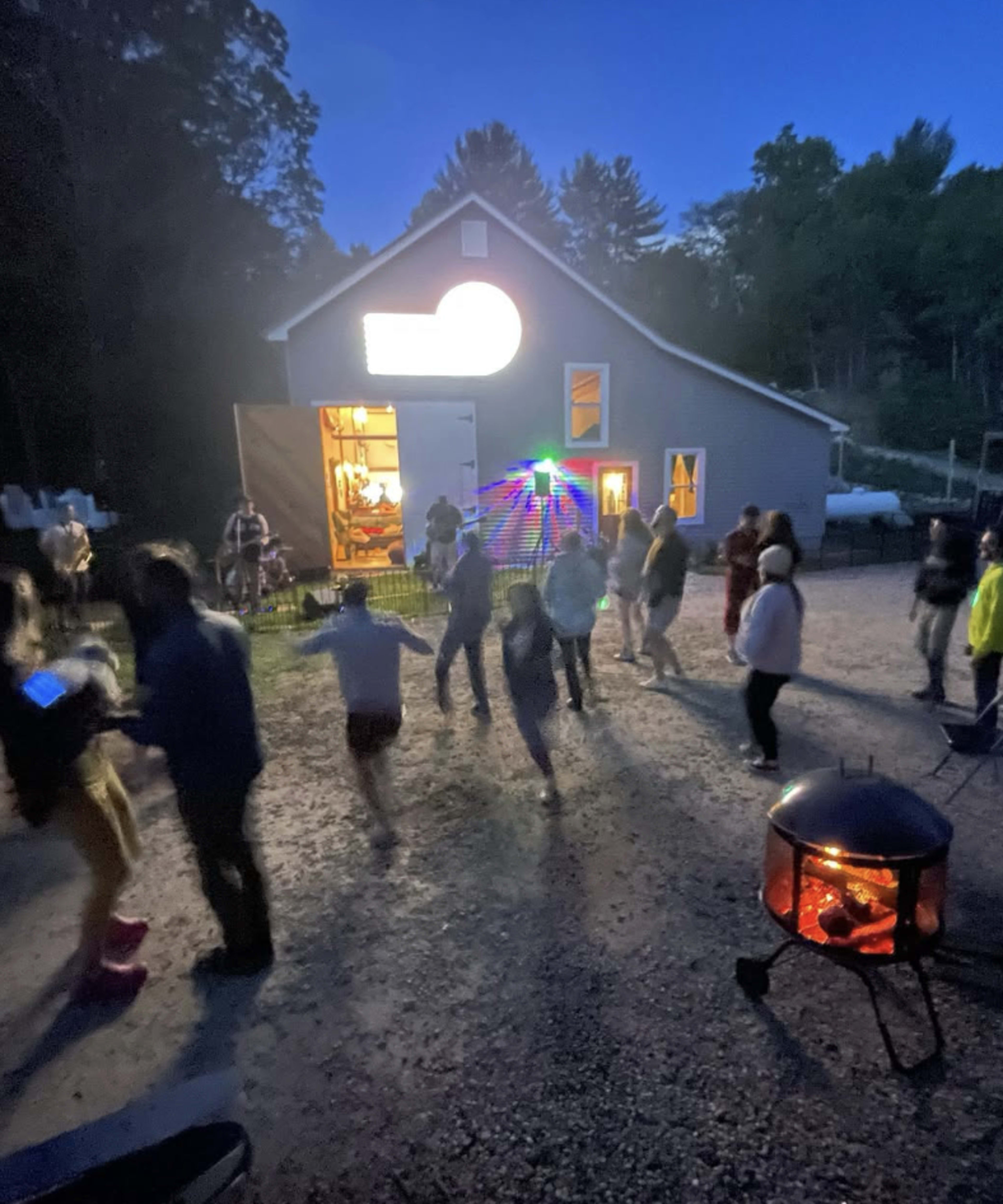 A group of people is dancing outside near a fire pit in front of a well-lit building during dusk.