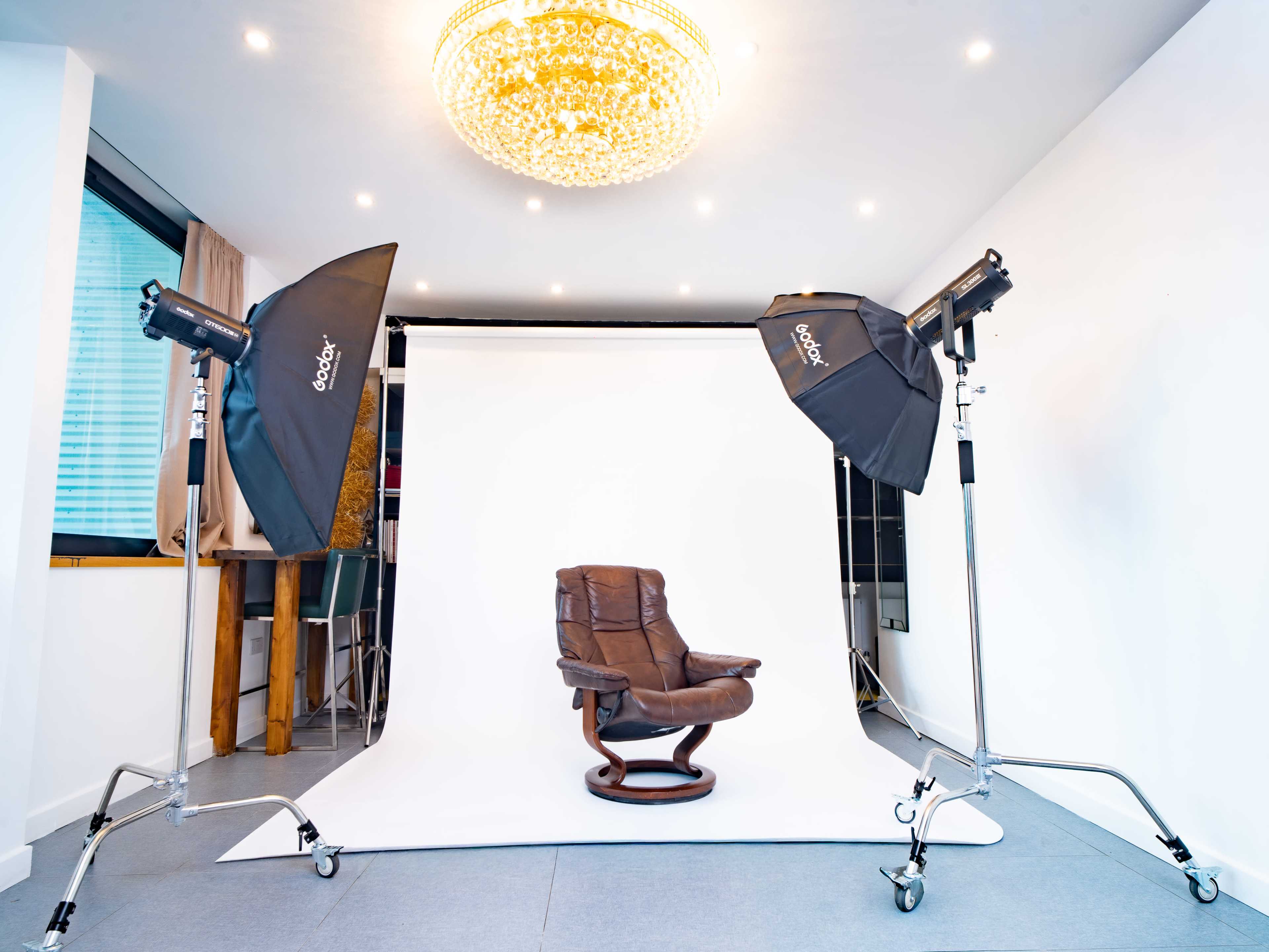 The image shows a photography studio with a brown reclining chair centered on a white backdrop, illuminated by two softbox lights and featuring a chandelier overhead.