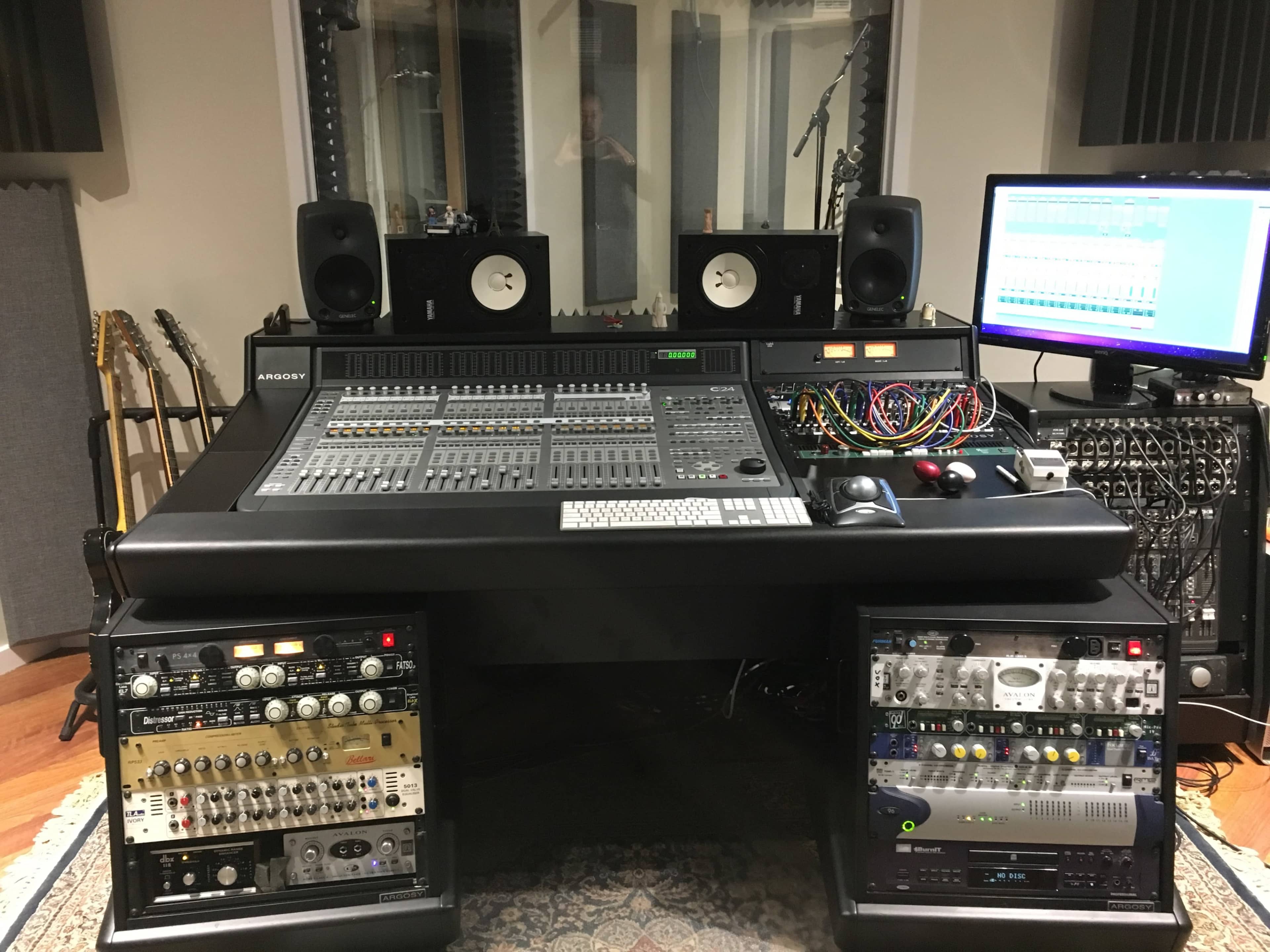 A professional audio mixing console surrounded by monitors, rack-mounted equipment, and a computer screen displaying audio tracks in a recording studio.