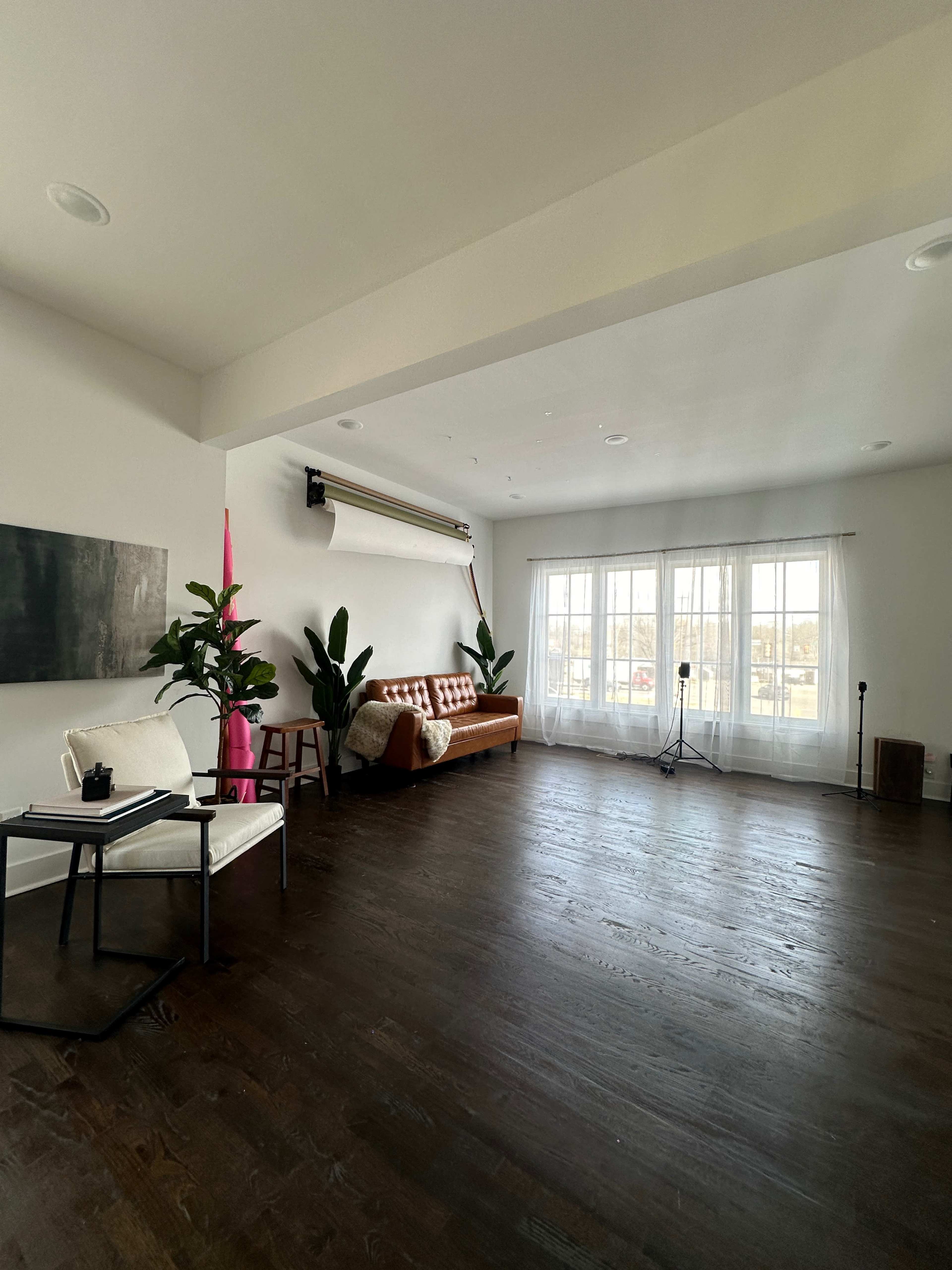 A spacious, well-lit room featuring a brown sofa, a white chair with a side table, and several potted plants near large windows.
