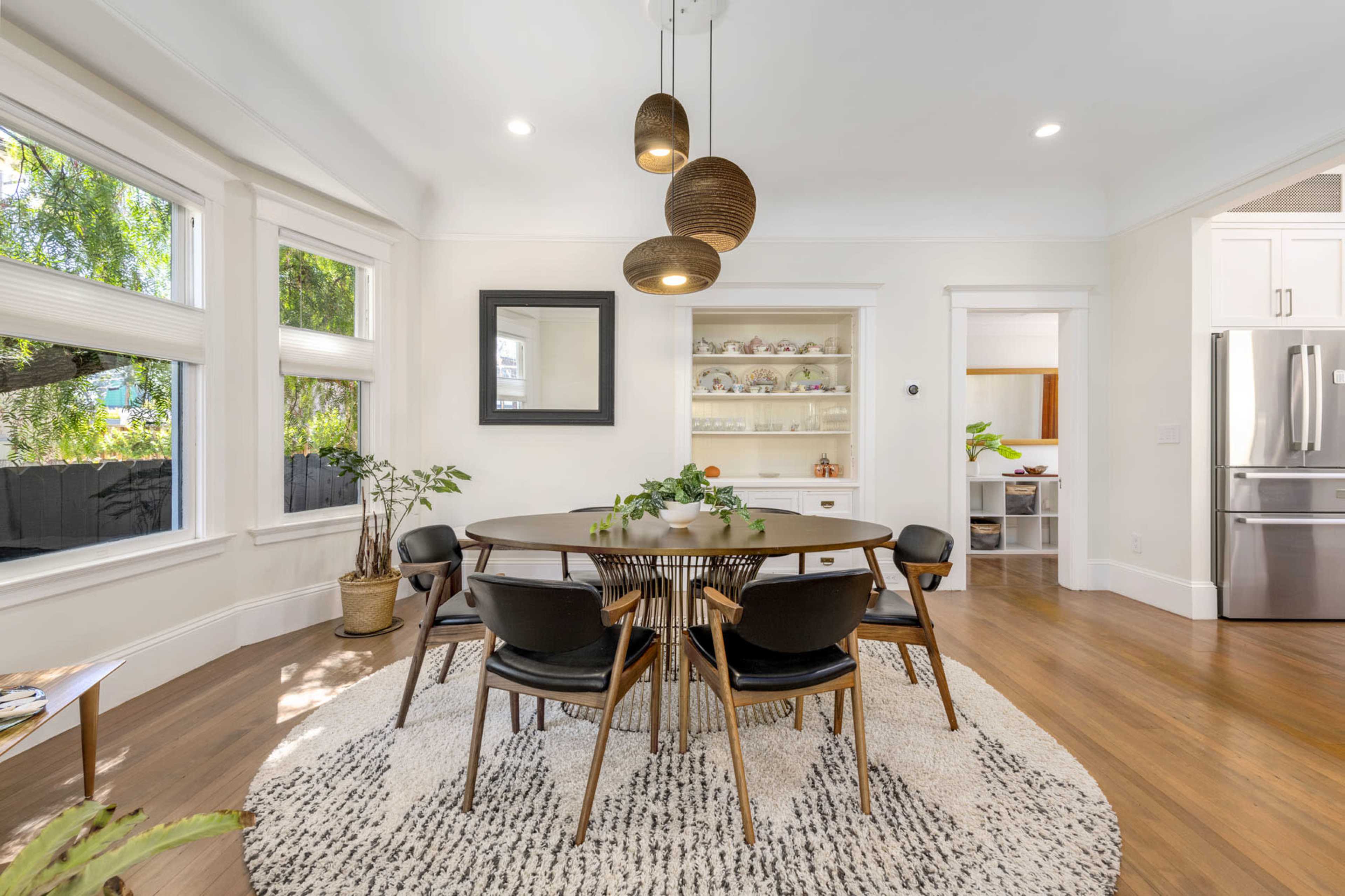A modern dining room features a round wooden table surrounded by black chairs, with pendant lights overhead and large windows letting in natural light.