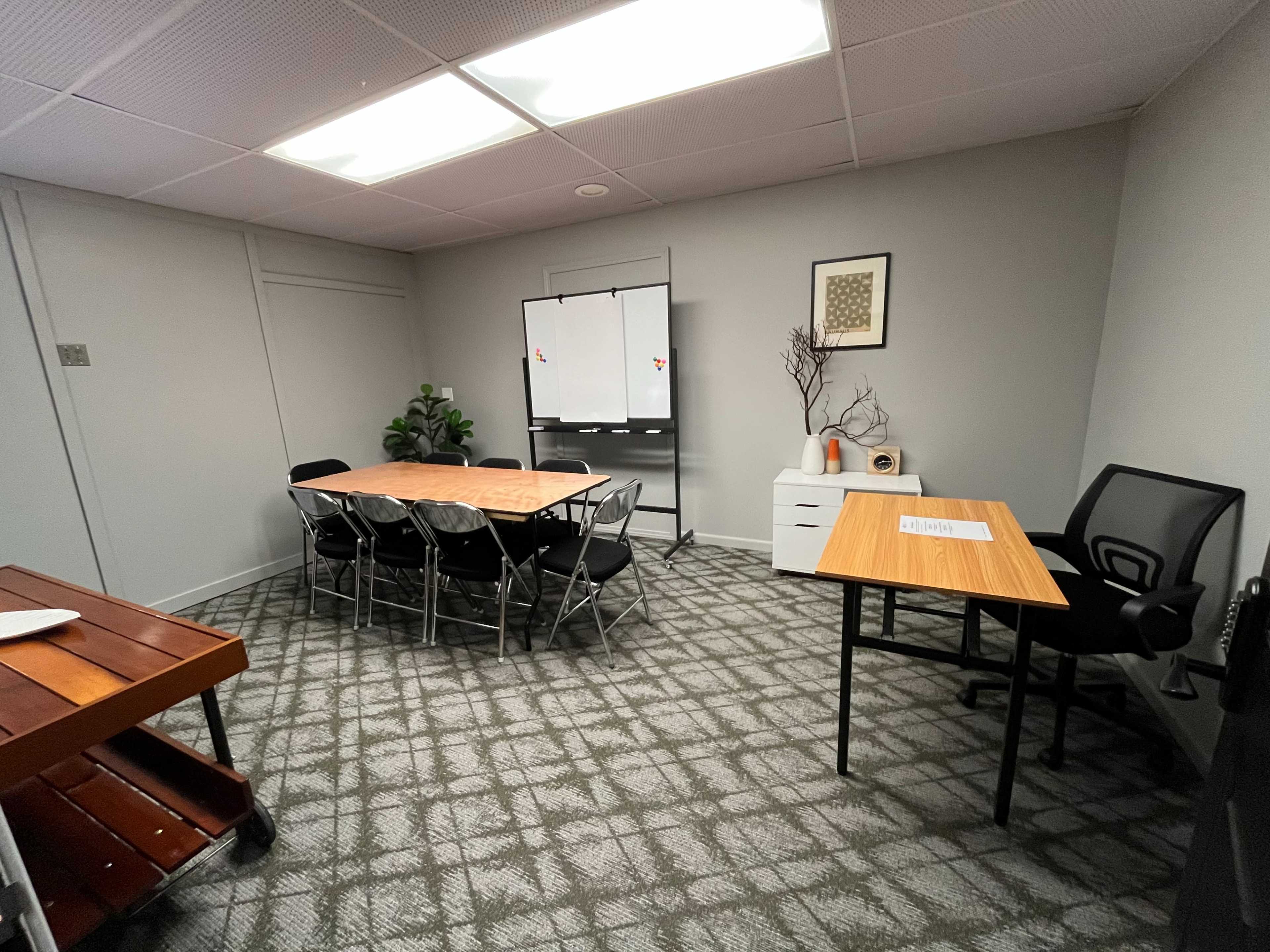 The image shows a meeting room with a rectangular table surrounded by chairs, a whiteboard on a stand, and a desk in one corner.