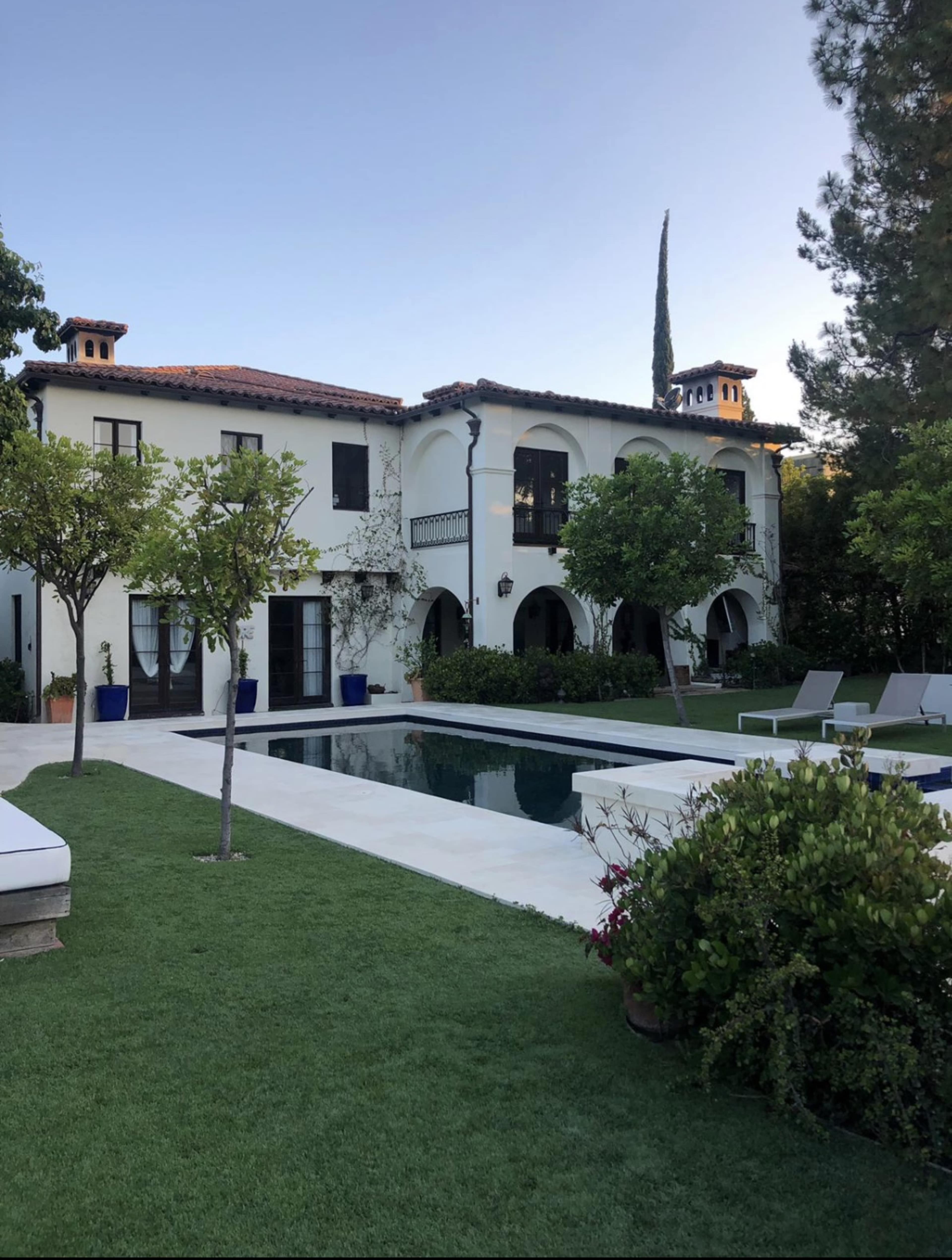 The image shows a two-story white villa surrounded by green lawns and trees, with a swimming pool in the foreground.