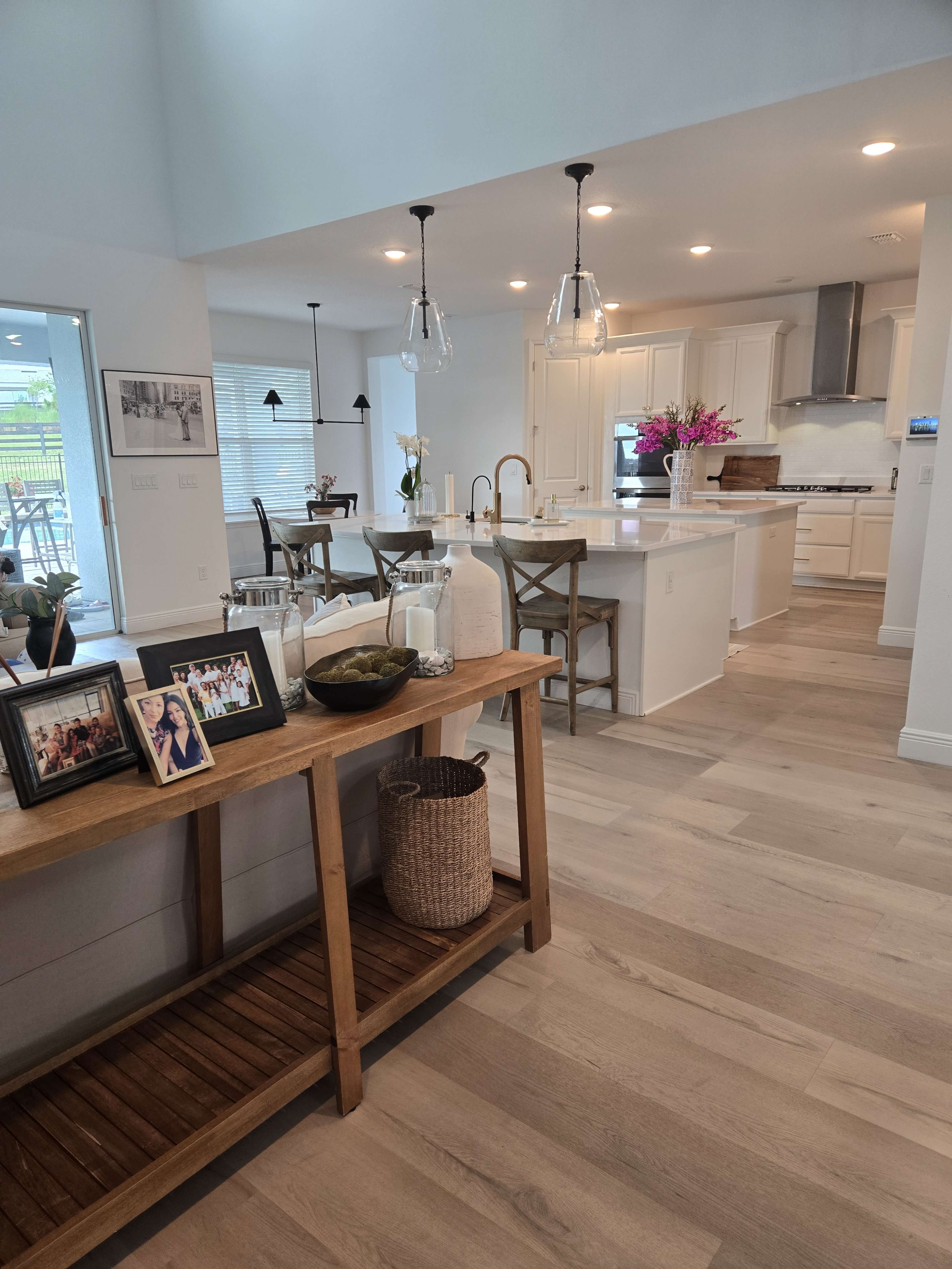 The image shows a modern open-concept kitchen and dining area with light-colored wood floors, pendant lighting, and a wooden console table displaying decor and framed photos.