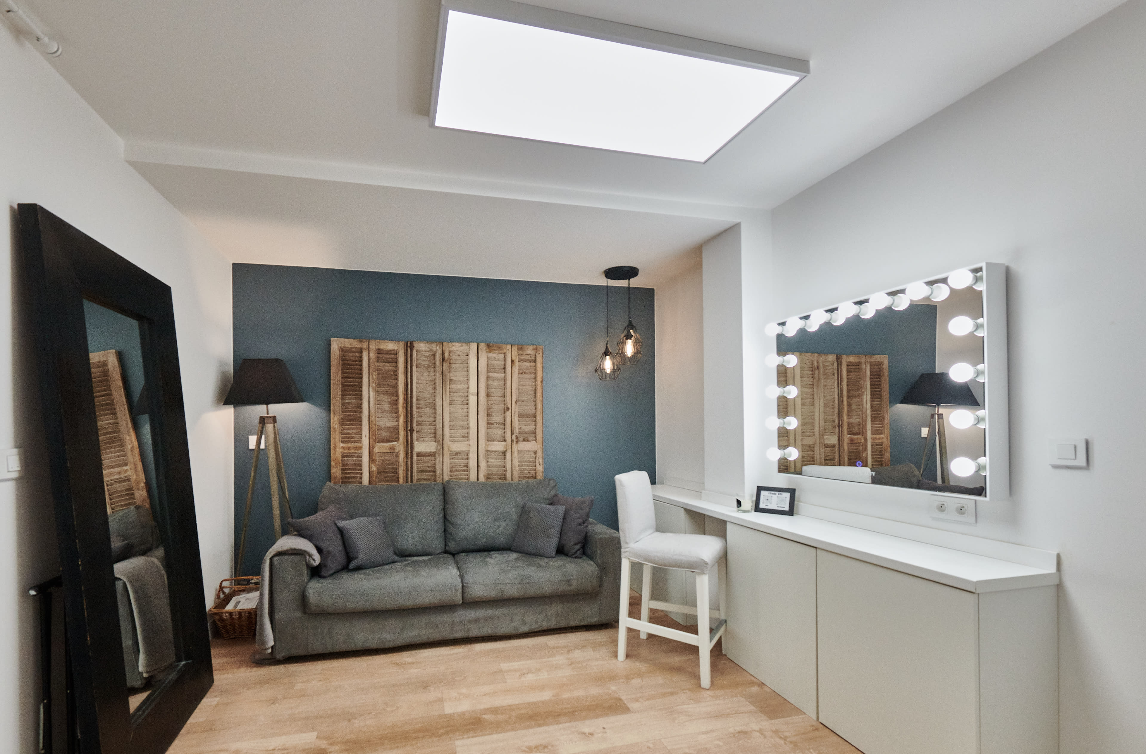 A room featuring a gray sofa, a large mirror with light bulbs, a white chair, and a wooden wall panel.