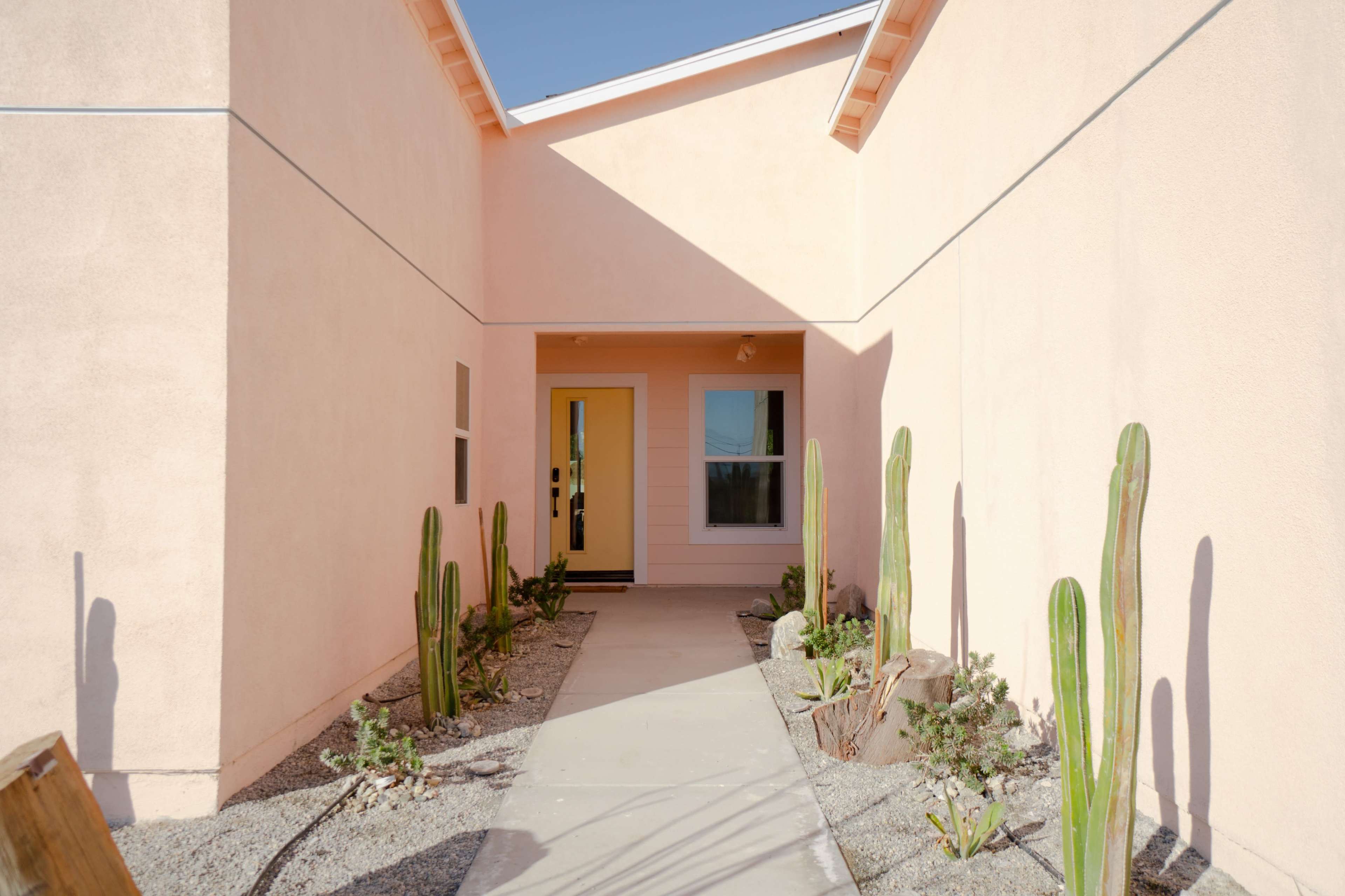 A pathway leading to a brightly painted house, flanked by several tall cacti and desert landscaping.