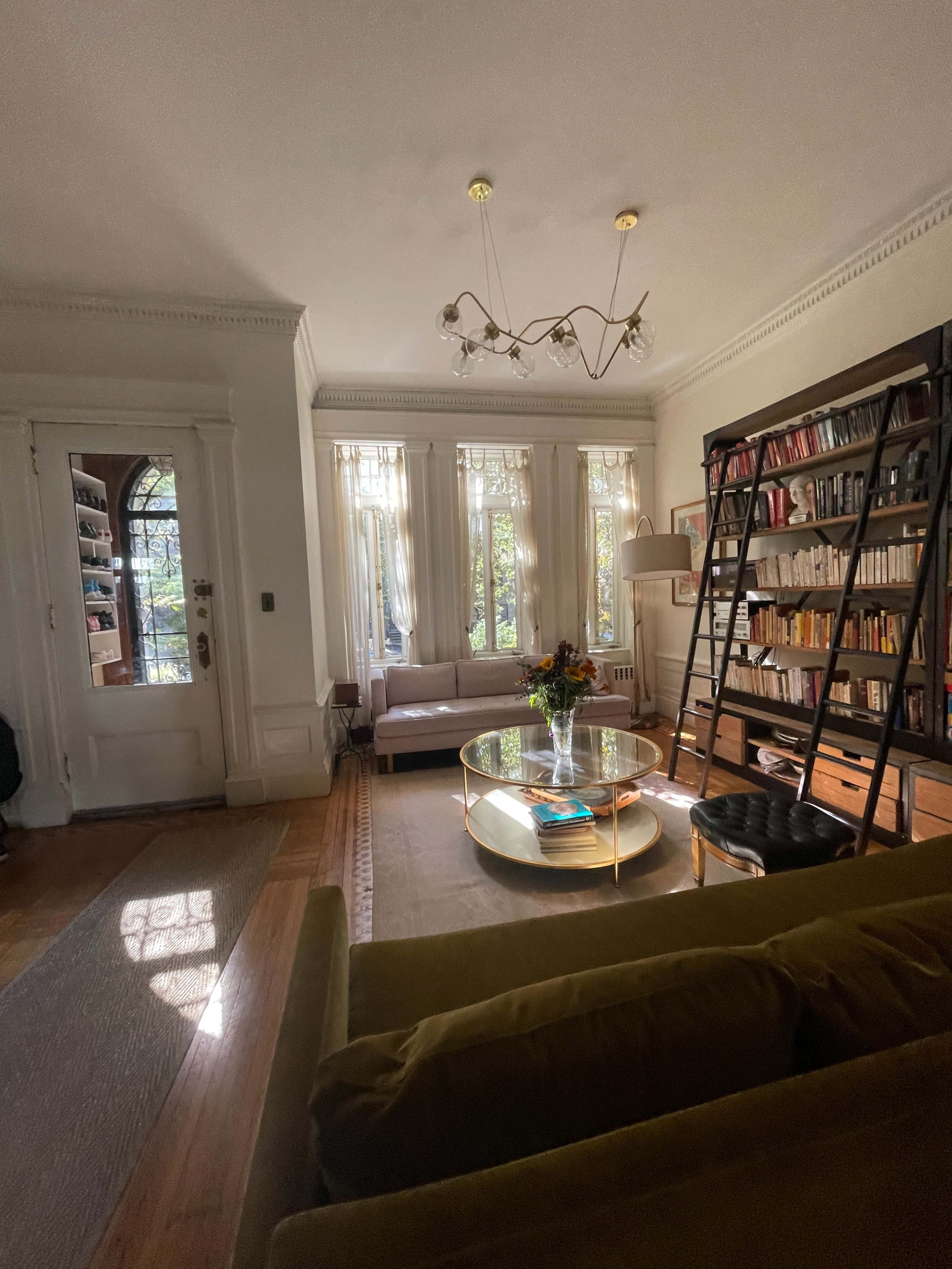 The image shows a cozy living room with a large bookshelf, a circular glass coffee table, and a green sofa, all illuminated by natural light coming through the windows.
