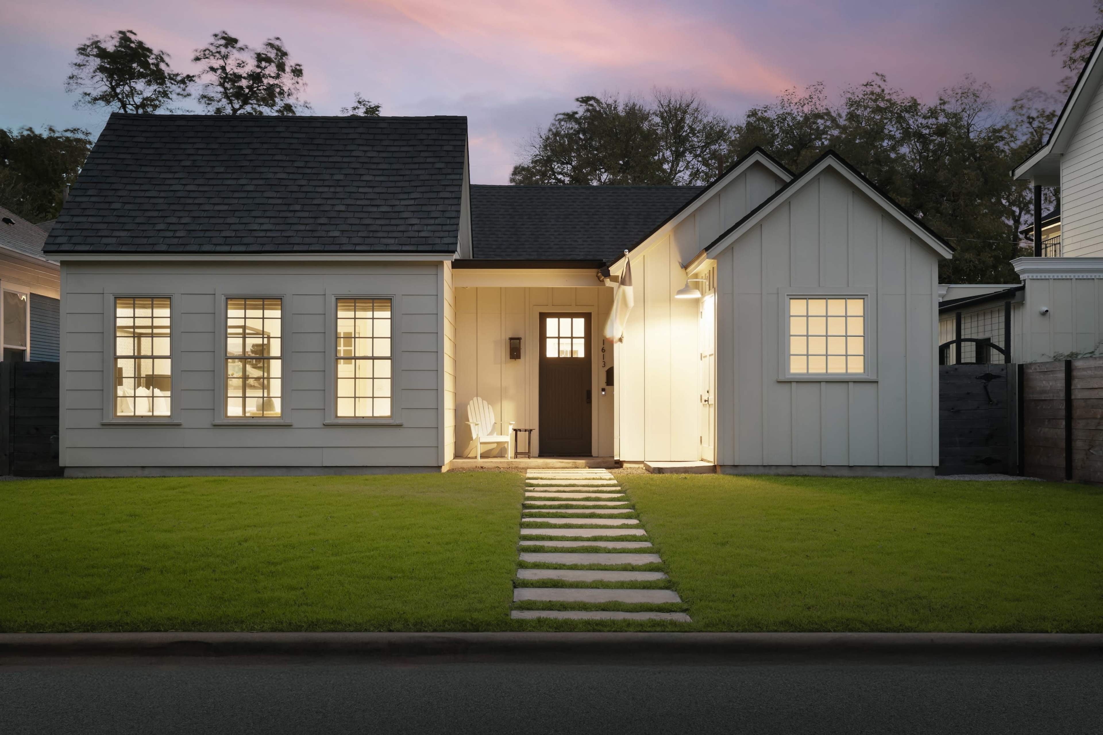 A modern one-story house with white clapboard siding and a black front door features illuminated windows and a stone pathway leading to the entrance, set against a twilight sky.
