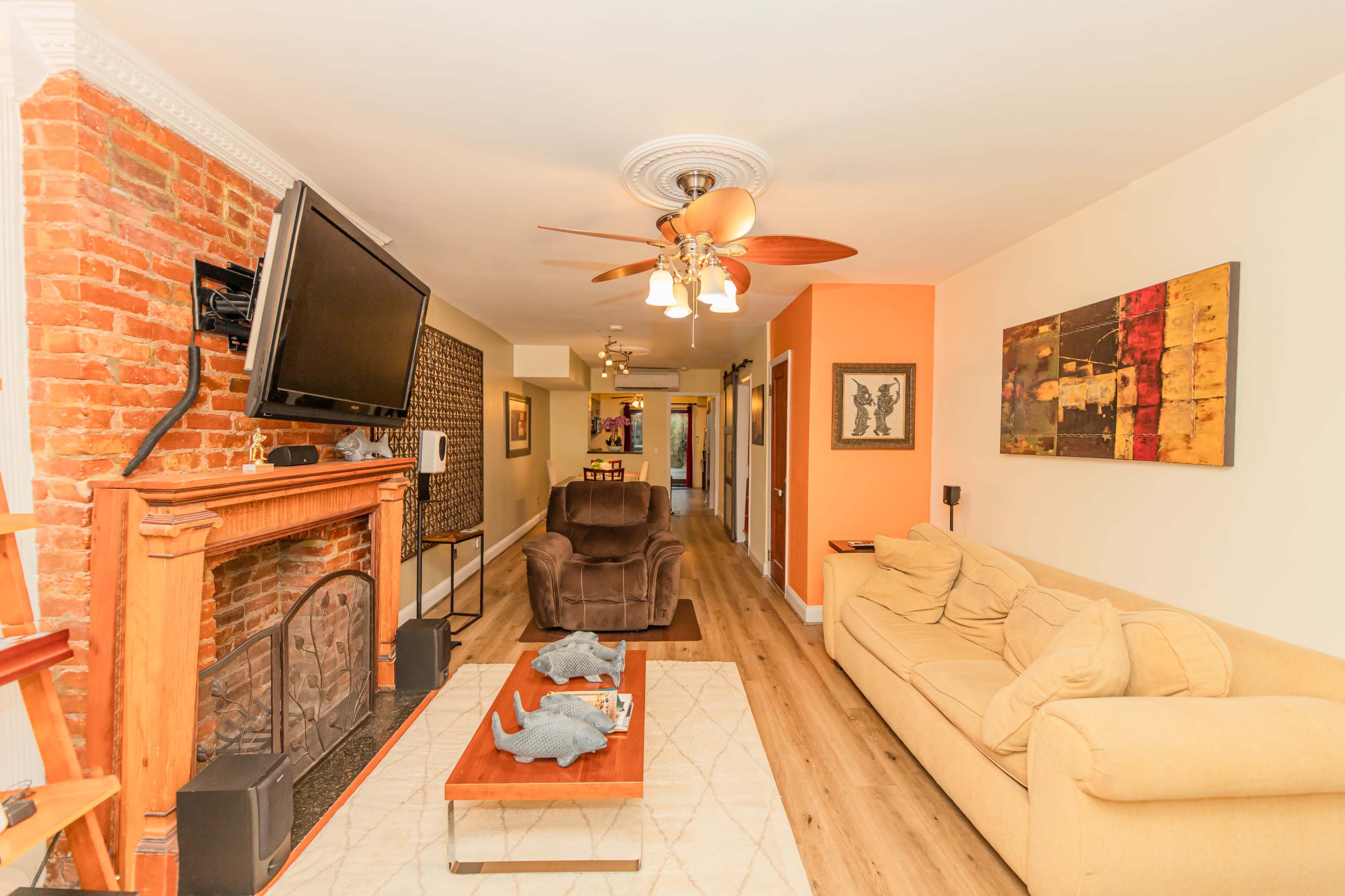 A living room with a brick fireplace, a flat-screen TV mounted on the wall, a beige sofa, and a wooden coffee table adorned with decorative items.