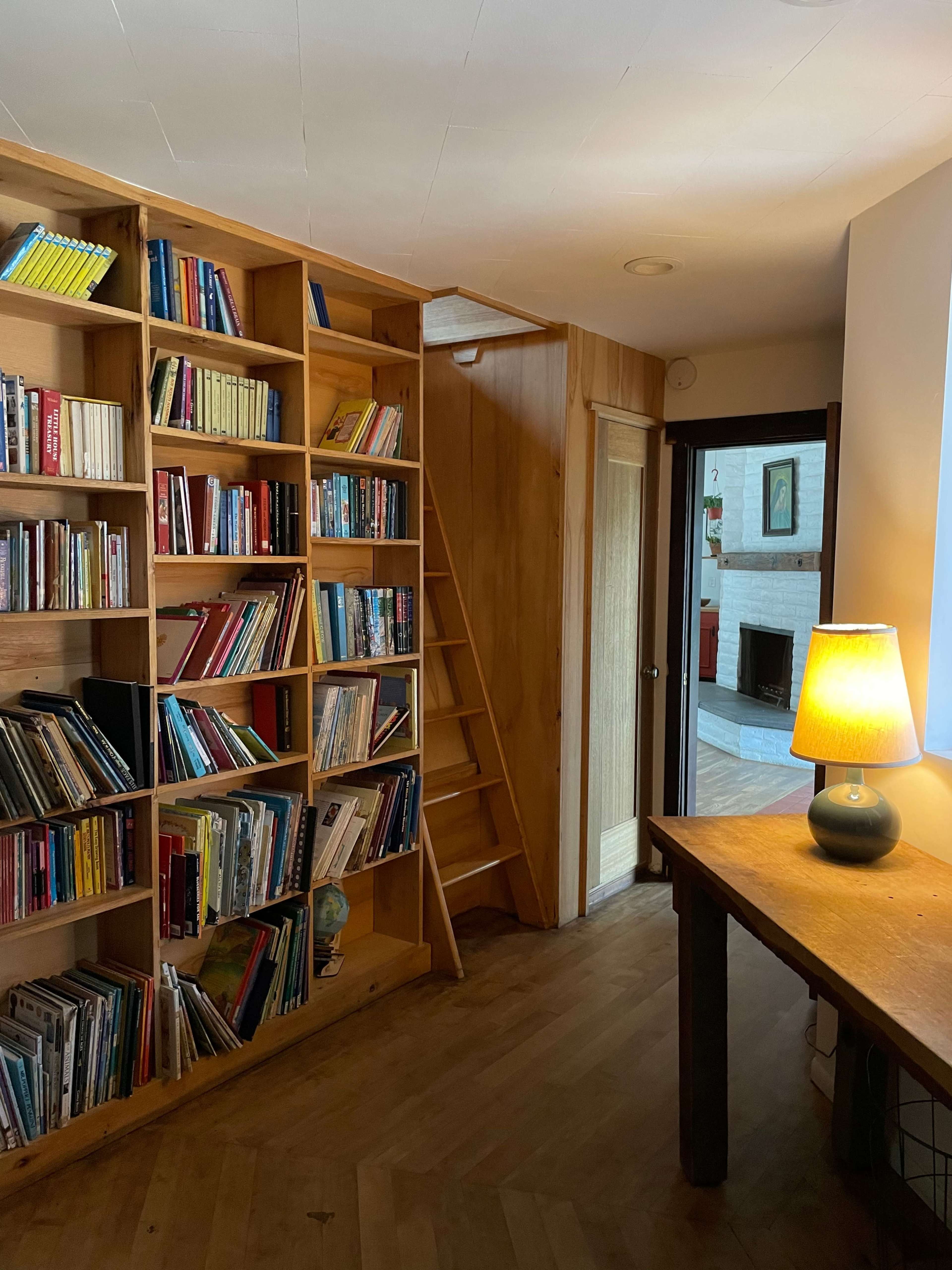 A cozy interior featuring a wooden bookshelf filled with books, a table with a lamp, and a staircase leading to another level.