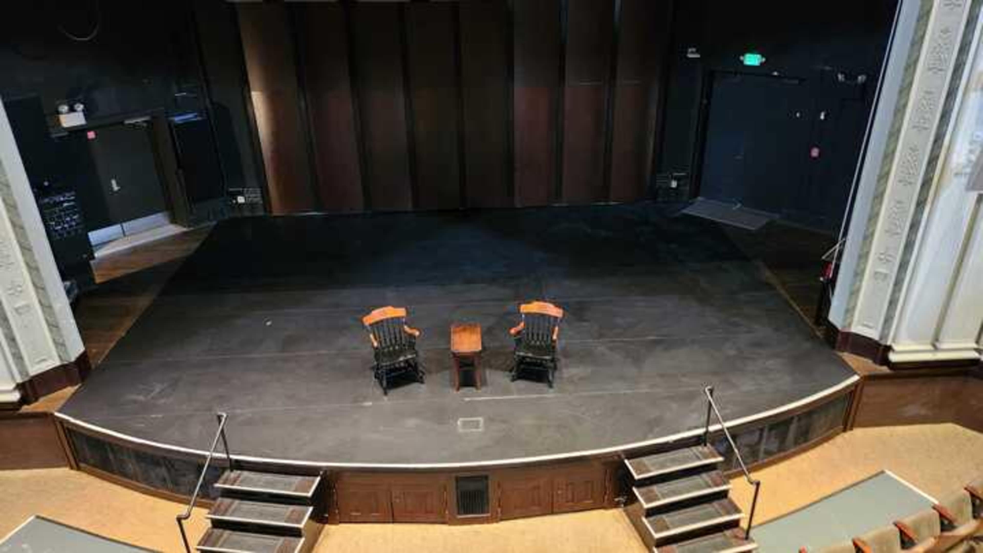 The image shows a staged theater with two wooden chairs and a small table placed on the blacked-out stage.