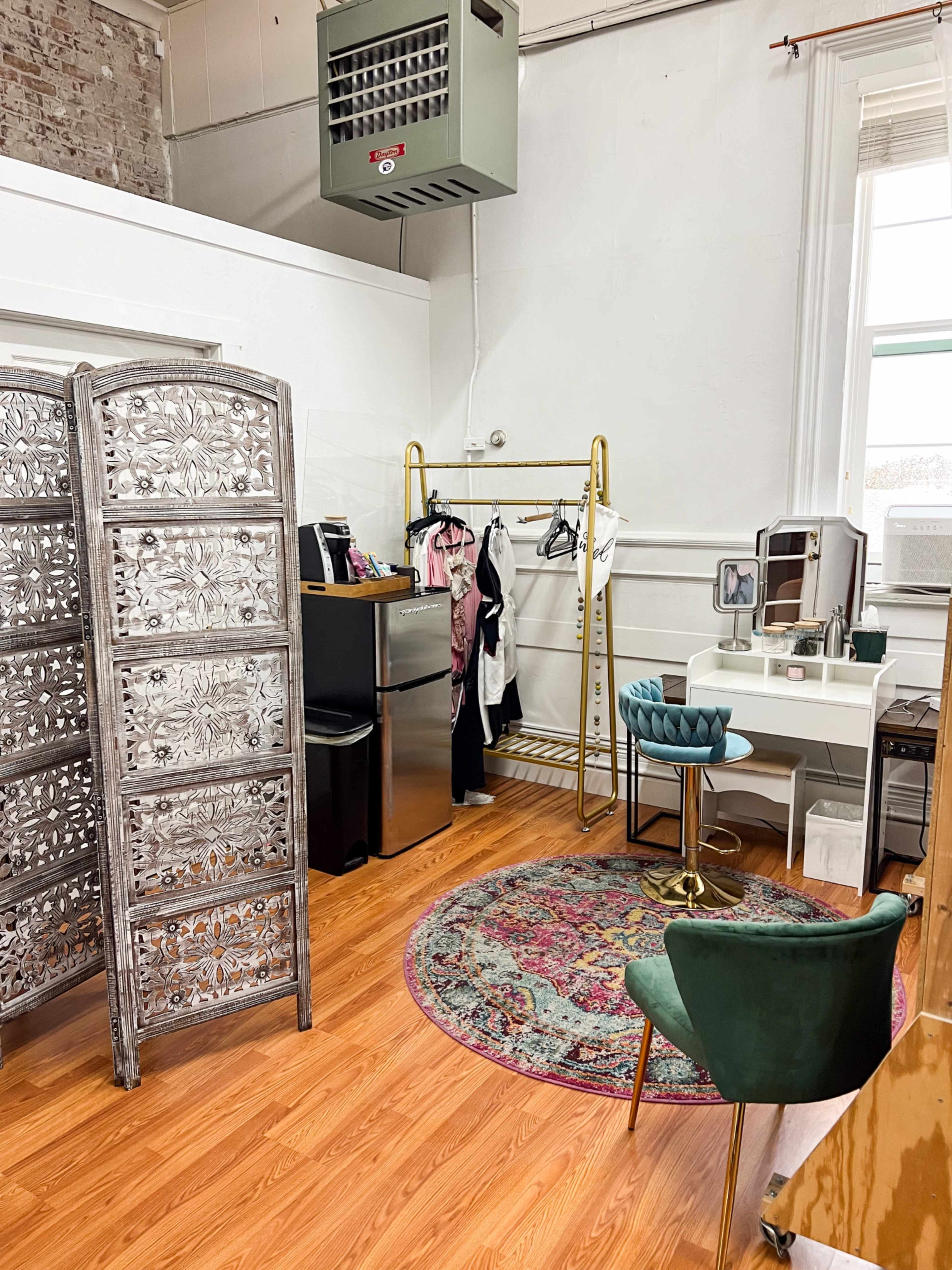 The image shows a room with wooden flooring featuring a decorative screen, a clothing rack, a small desk with a chair, a mini refrigerator, and an air conditioning unit.