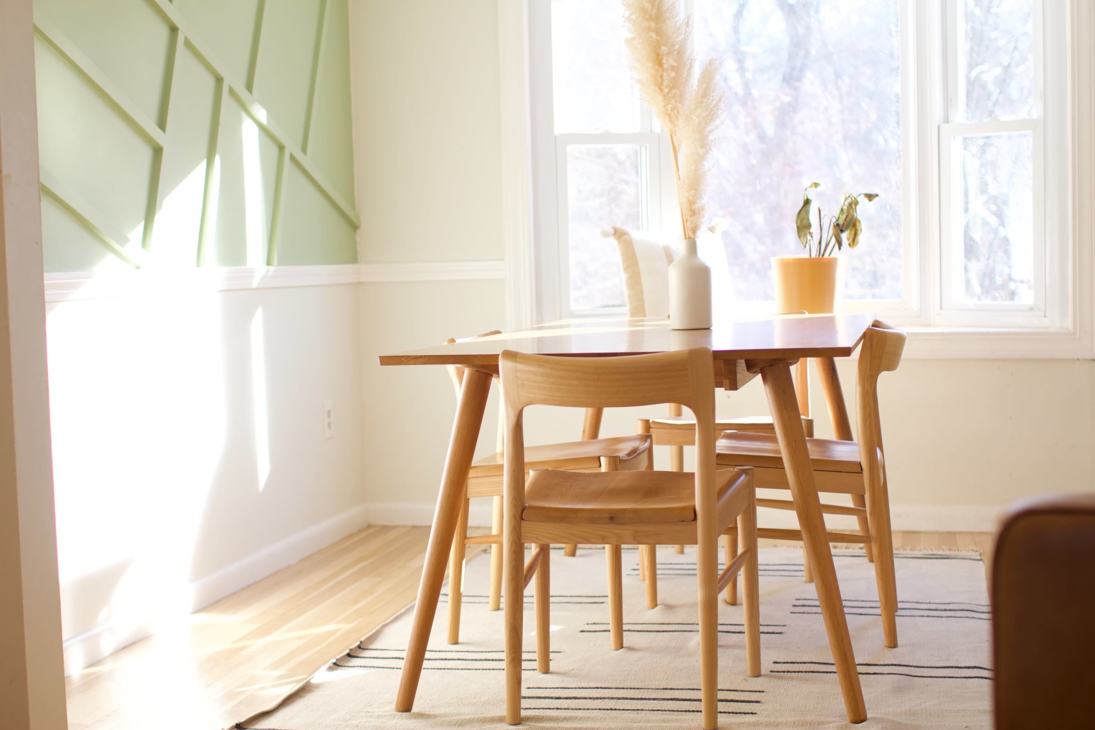A wooden dining table with three chairs is positioned on a patterned rug in a well-lit room featuring green wall accents and a window.