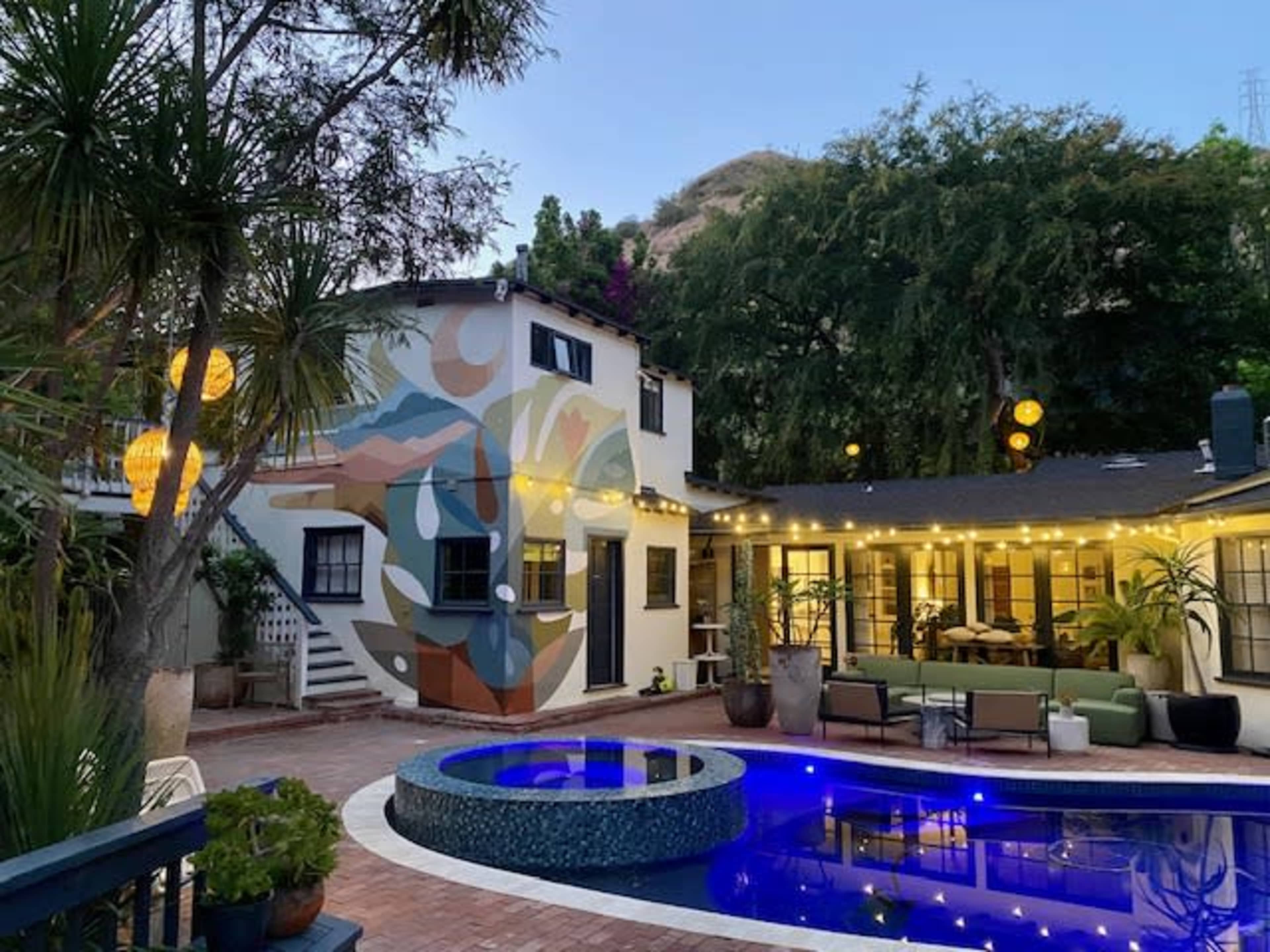 The image shows a backyard with a pool, surrounded by a colorful mural on a building, string lights hanging above, and a seating area with greenery.