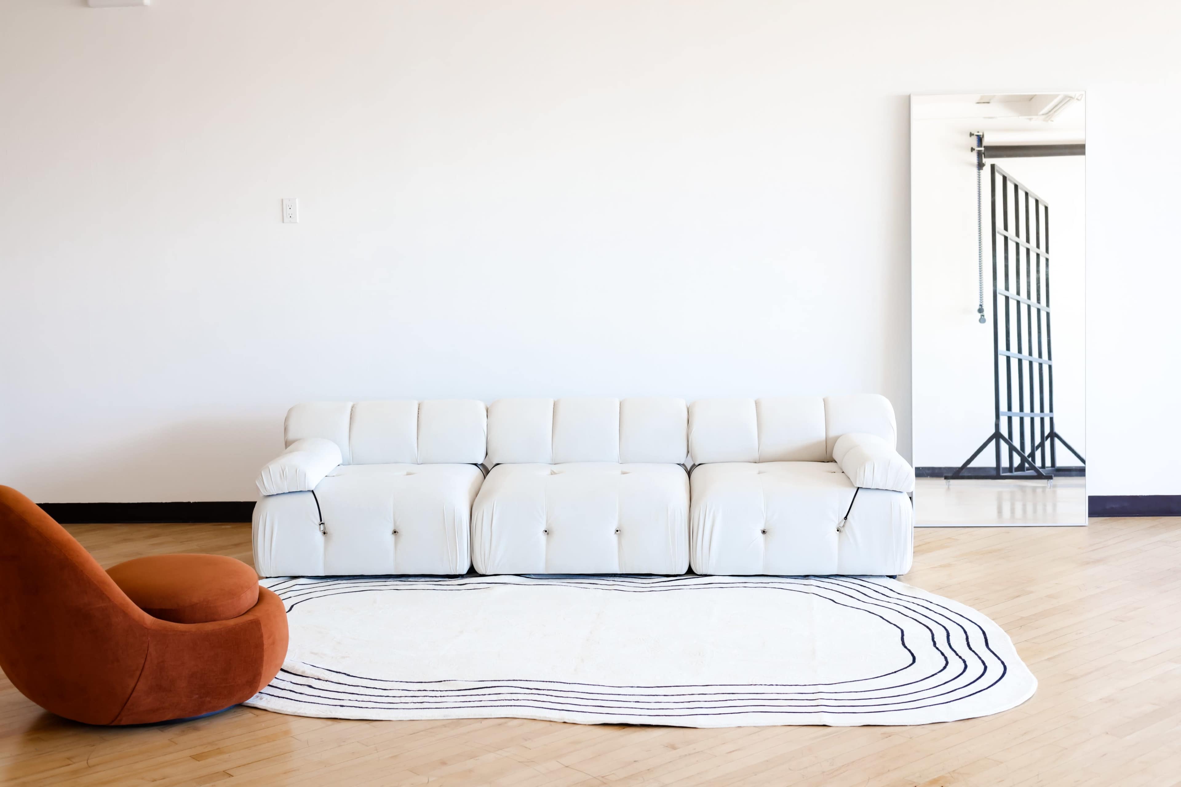 A white sofa and an orange chair are positioned in a minimalistic room with a large mirror on the wall.
