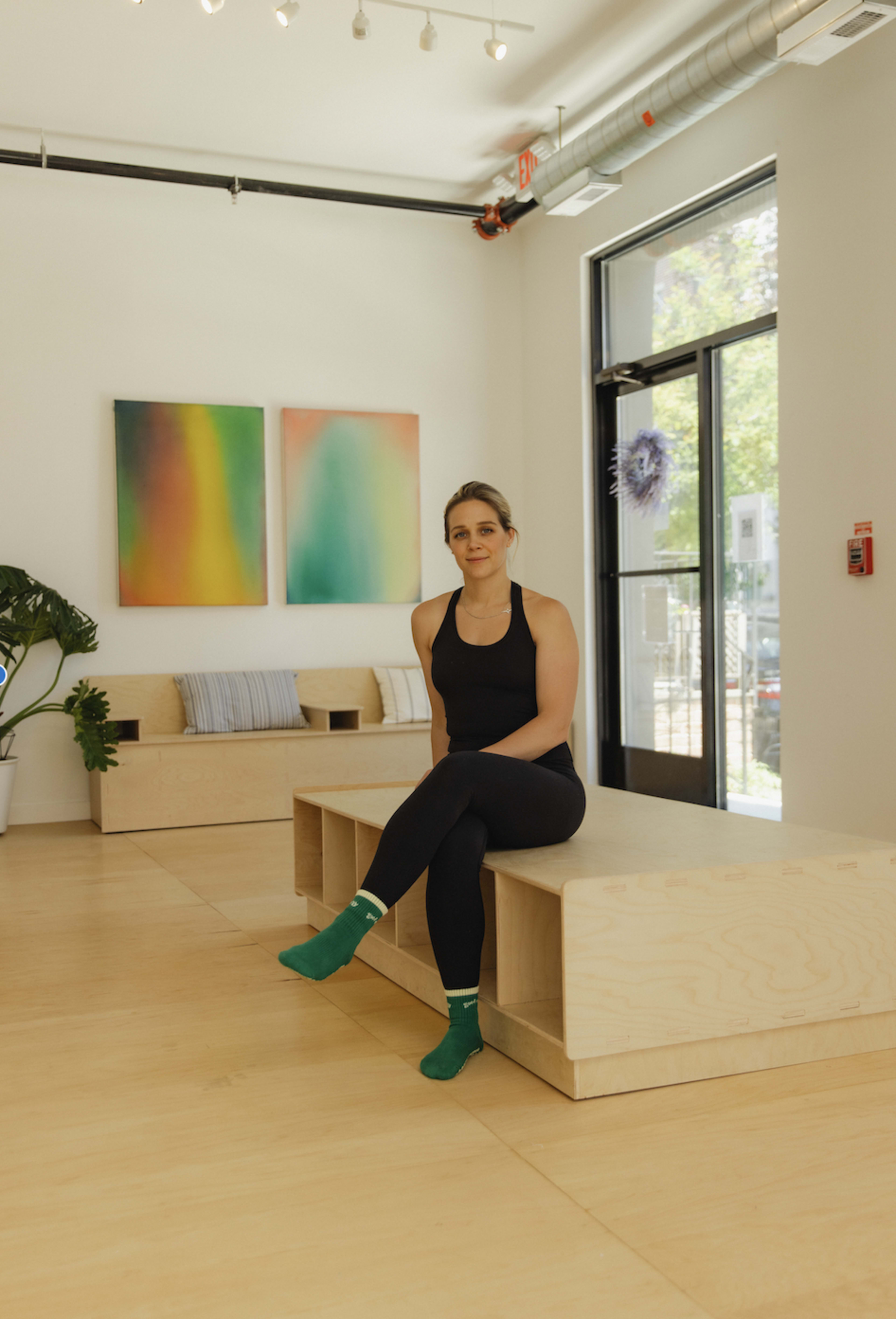 A woman in black athletic wear sits on a wooden bench in a bright, minimalist room with large windows and abstract paintings on the wall.
