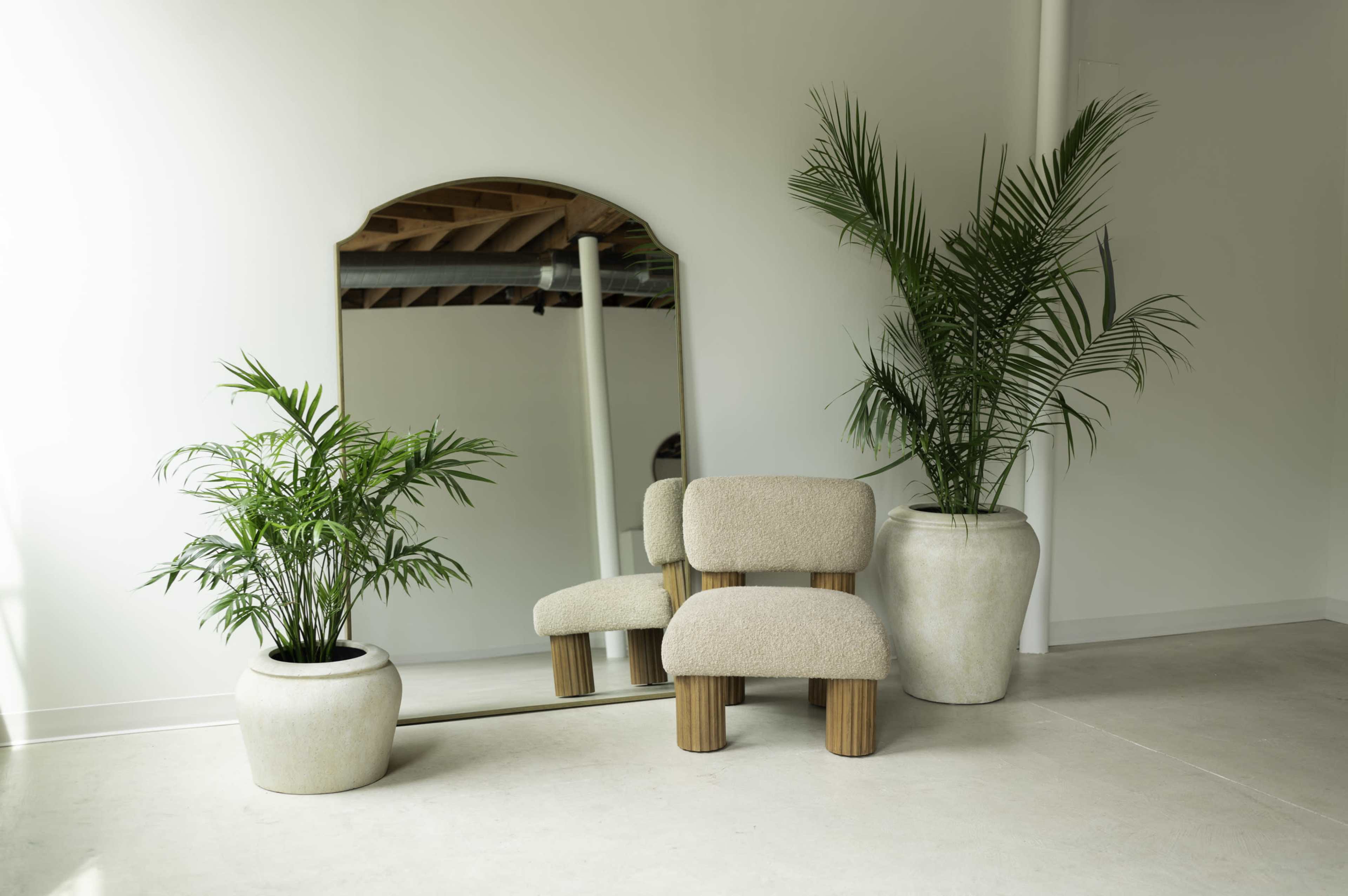 The image features a minimalistic interior with a textured chair, a large mirror, and two potted plants beside a concrete floor.
