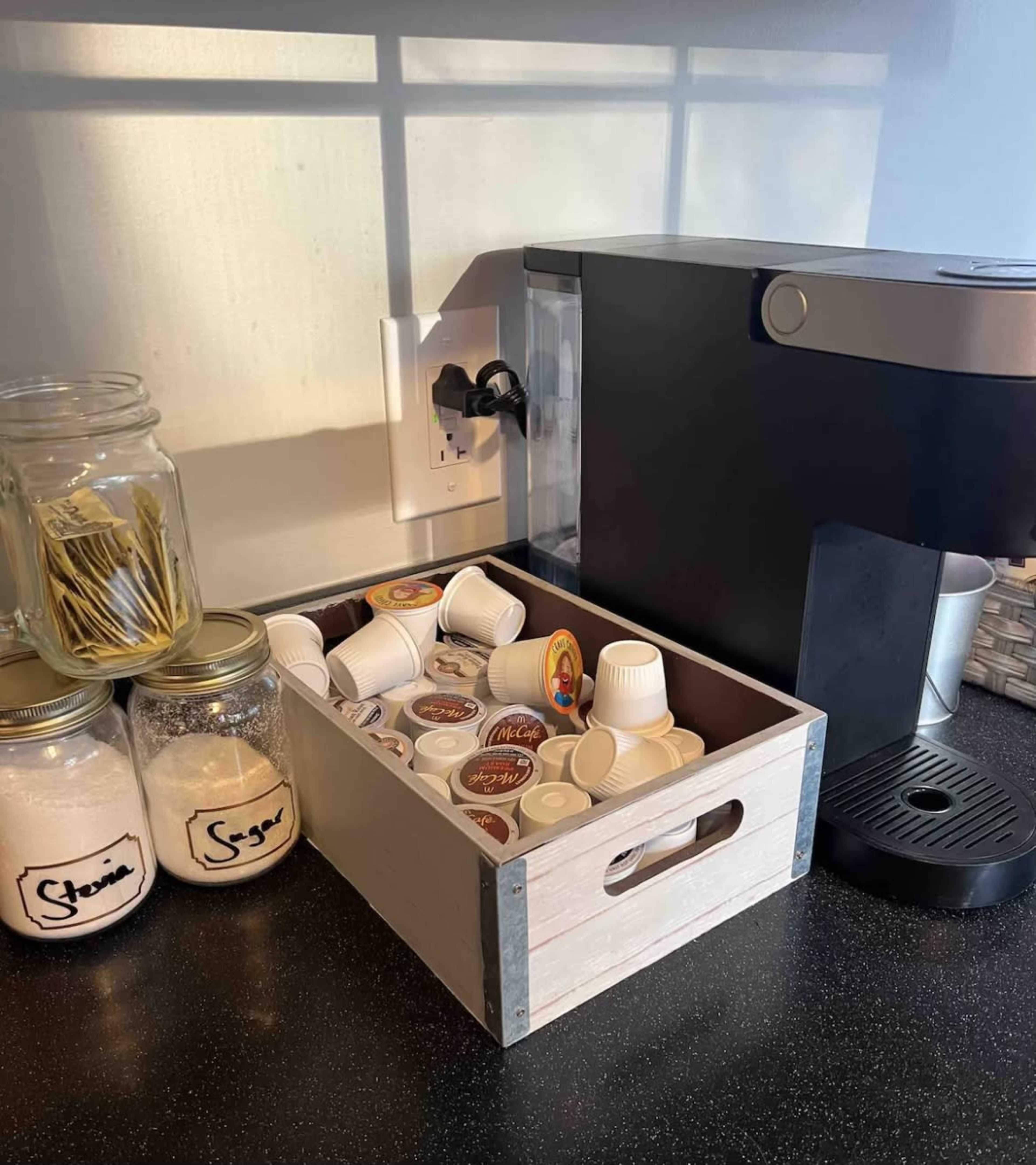 A wooden box filled with coffee pods sits next to a coffee machine on a countertop, alongside jars labeled "Stevia" and "Sugar."