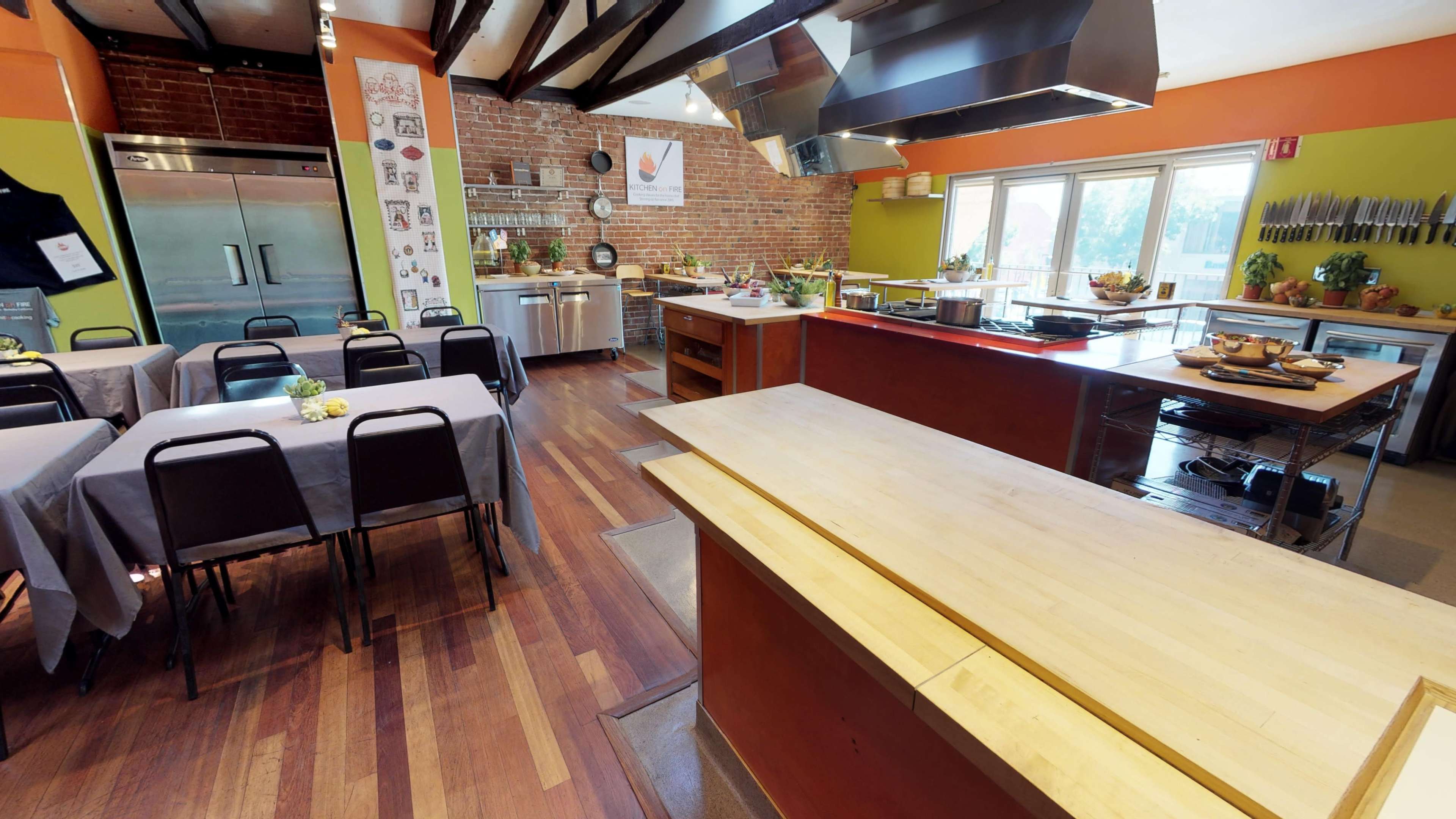 The image shows a spacious kitchen with brick walls, wooden flooring, and multiple cooking stations, along with tables and chairs arranged for dining.